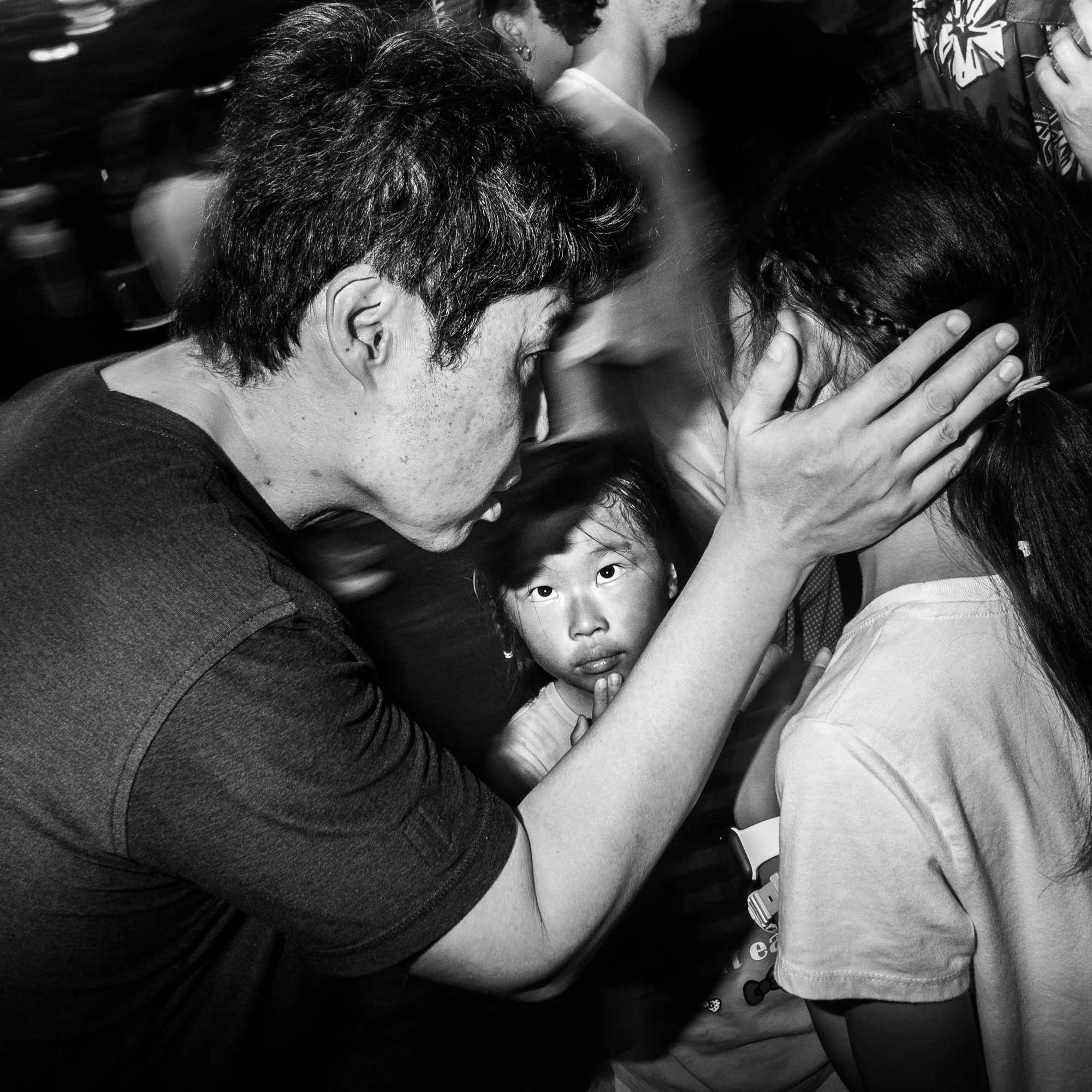 A black-and-white photo of a young girl with long dark hair, and an older woman with short dark hair, who are kissing each other on the face. The girl is wearing a light-colored shirt, and the woman has her eyes closed, with her hand gently placed on