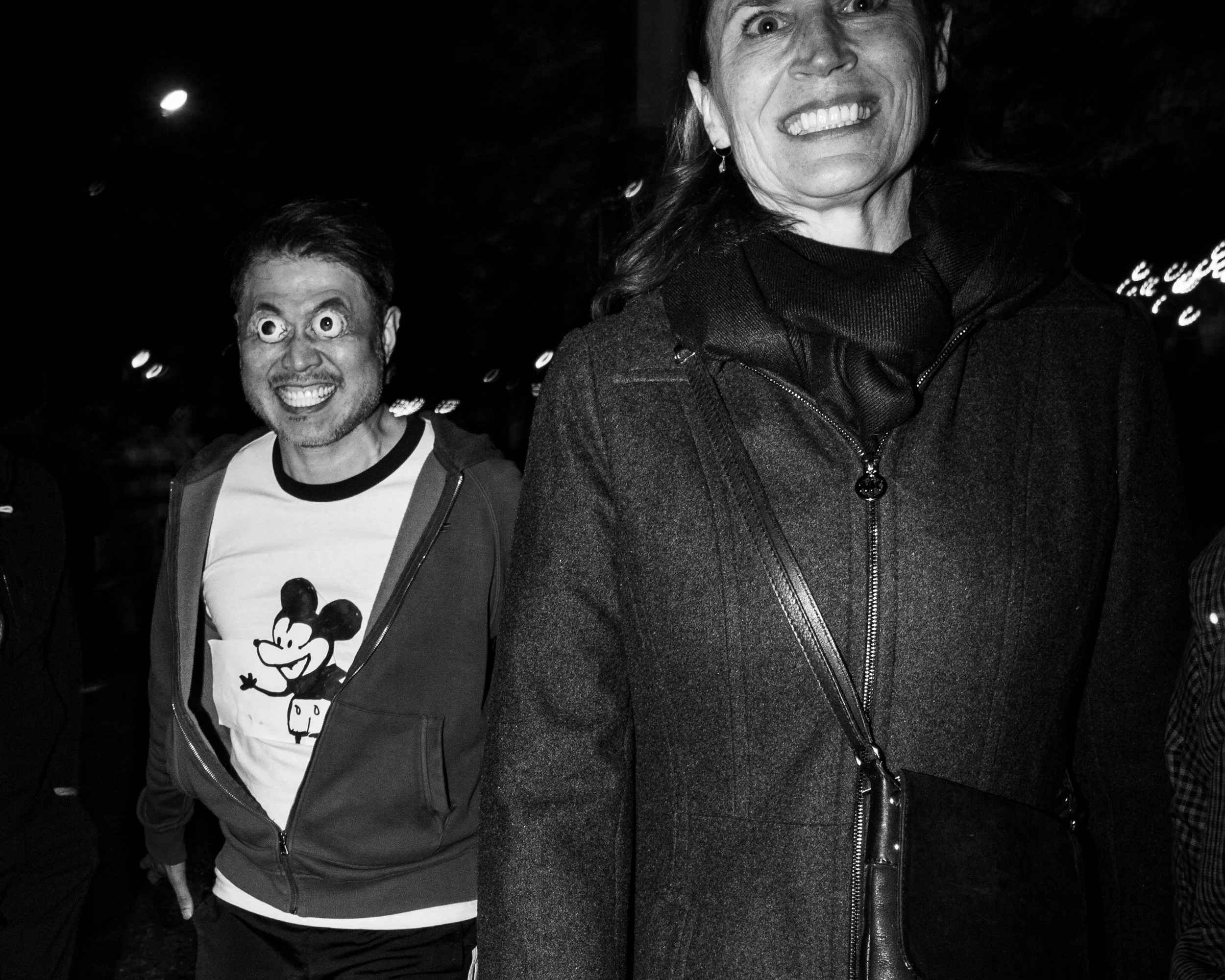 Two people smiling at night; the man on the left wearing glasses and a shirt with Mickey Mouse, and the woman on the right wearing a dark jacket and earrings, both with wide smiles.