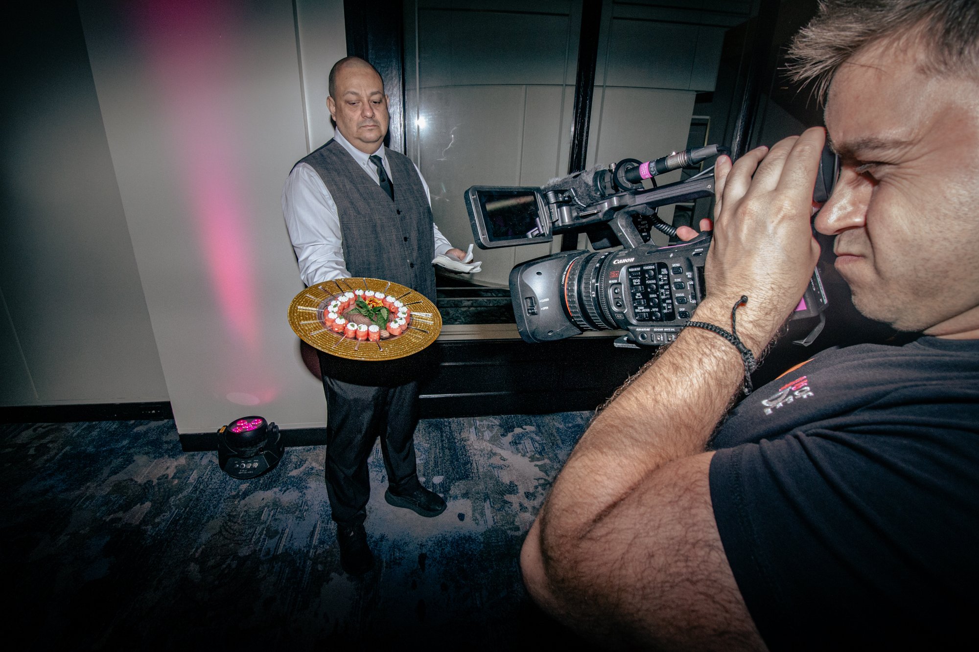 A man is holding a professional video camera and filming a man in formal attire who is holding a plate of sushi in an indoor setting.