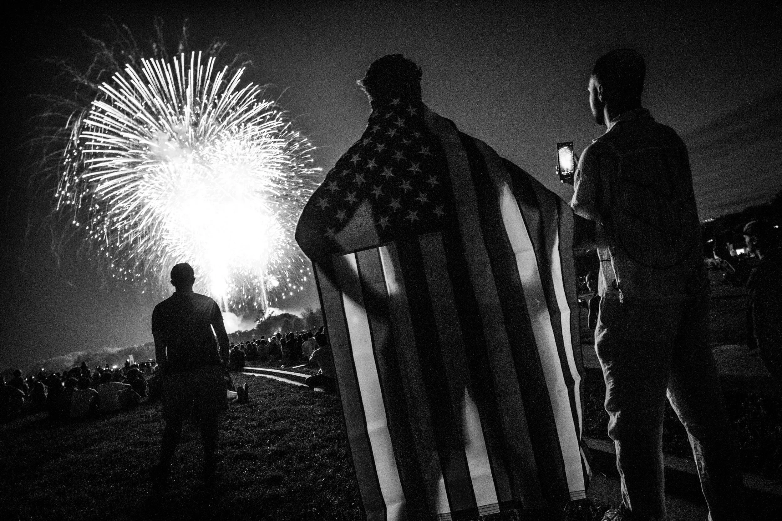 Black and white photo of people watching fireworks, two individuals are standing near a flag-draped box or speaker, with one holding a phone, with fireworks lighting up the sky in the background.