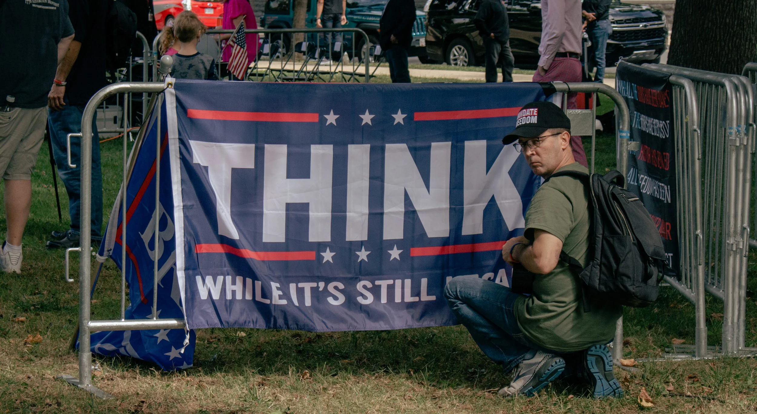 A man with glasses kneeling next to a large Trump flag at a political gathering. Several people are standing around, and parked cars are visible in the background.