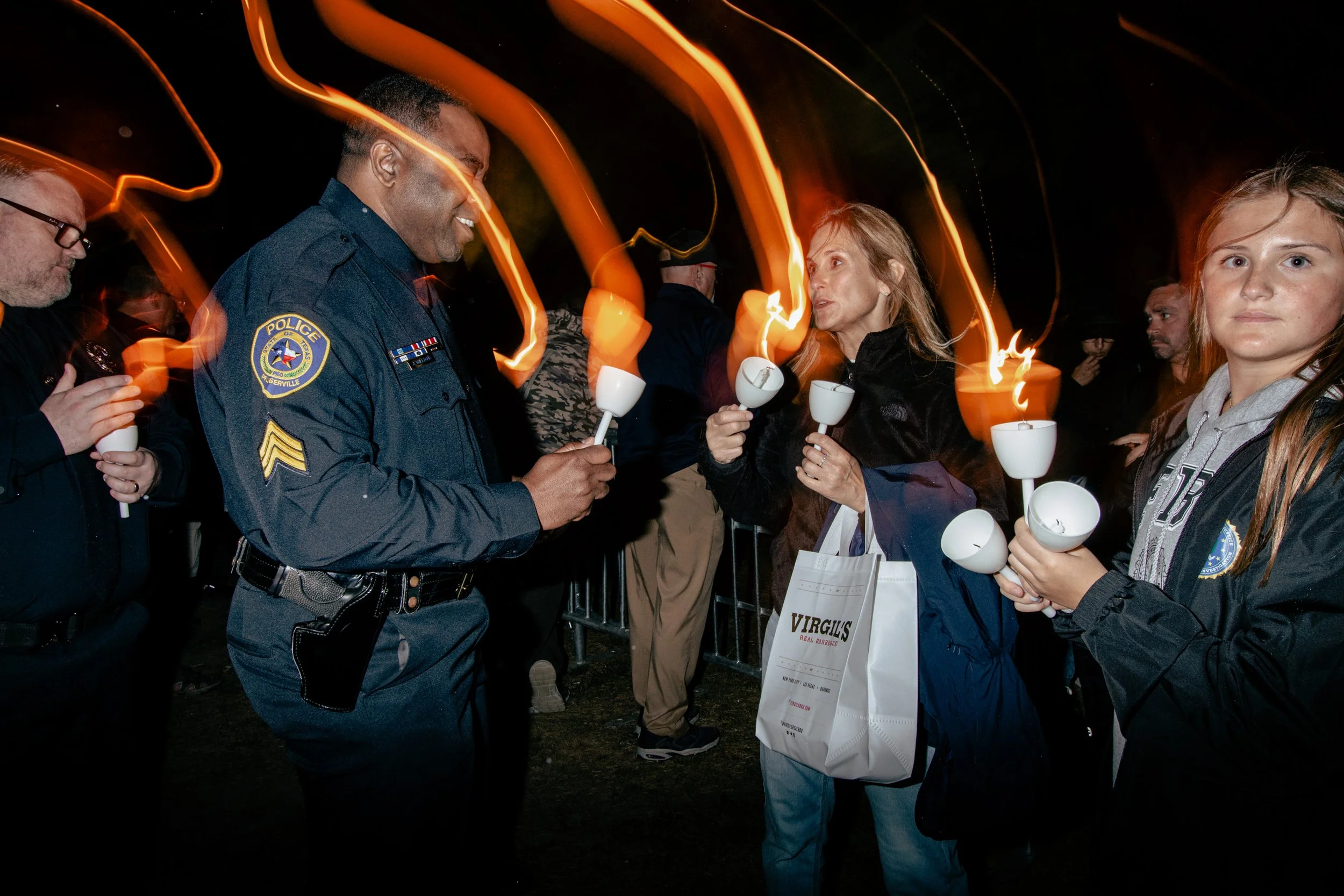 Police officers and civilians gathered at night holding white candles during a memorial or vigil, with orange flame-like light streaks in the background.