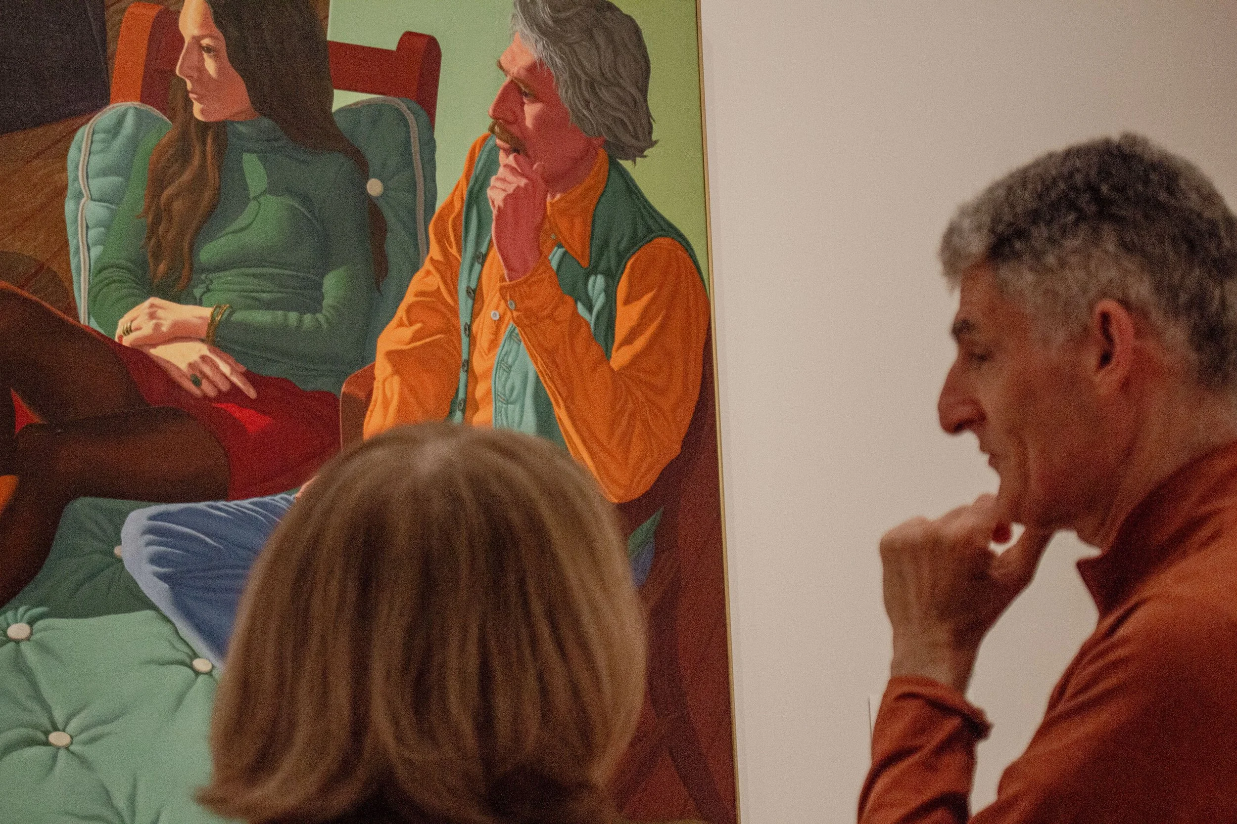 A man with gray hair and a mustache, wearing an orange shirt and dark green vest, sitting with his hand on his chin, appears to be contemplating. To his left, a woman with long dark hair, wearing a green jacket, is seated with her hands folded on her