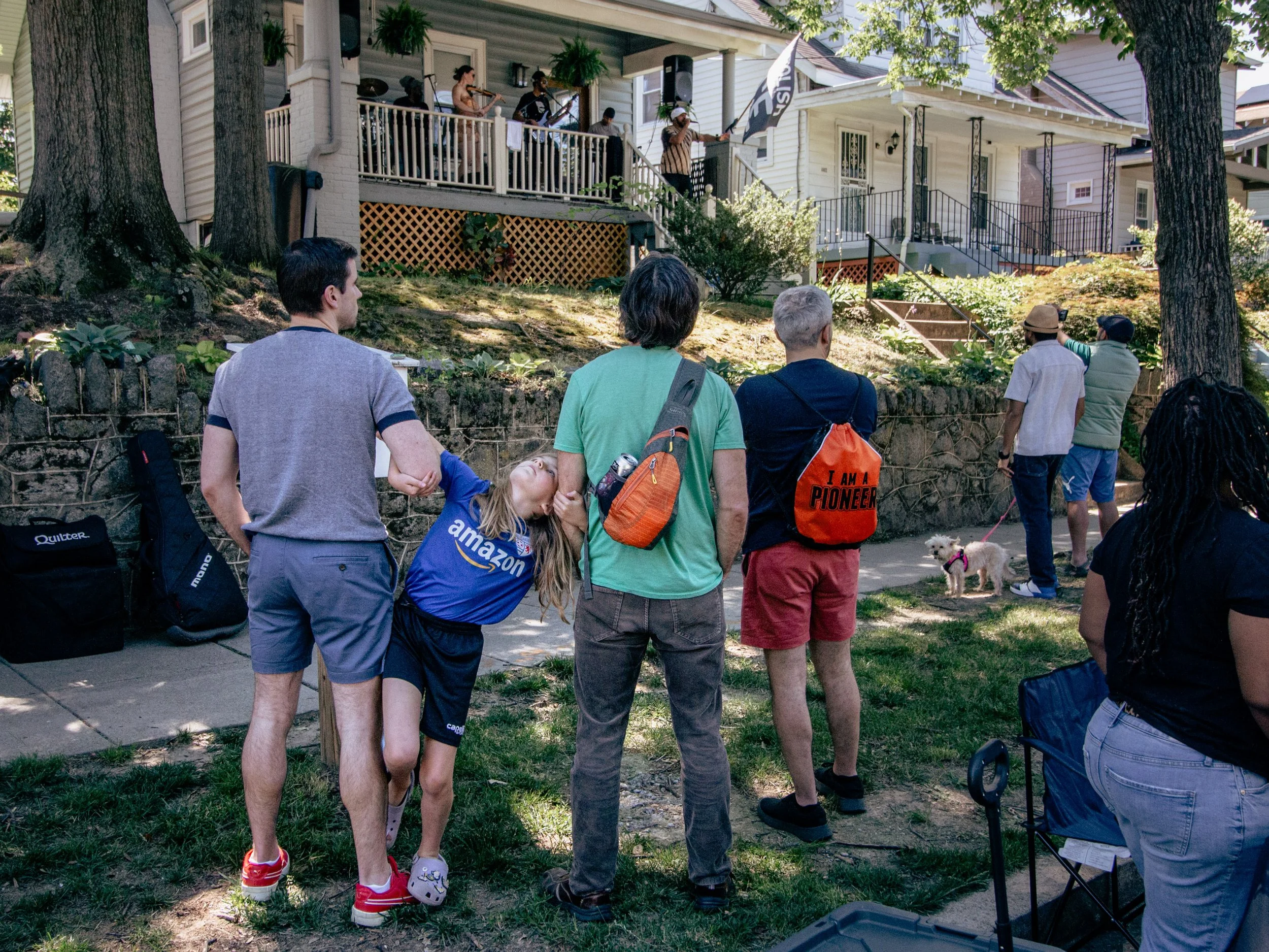 People gathered outdoors watching a band perform on a house porch, with trees and greenery surrounding the scene.