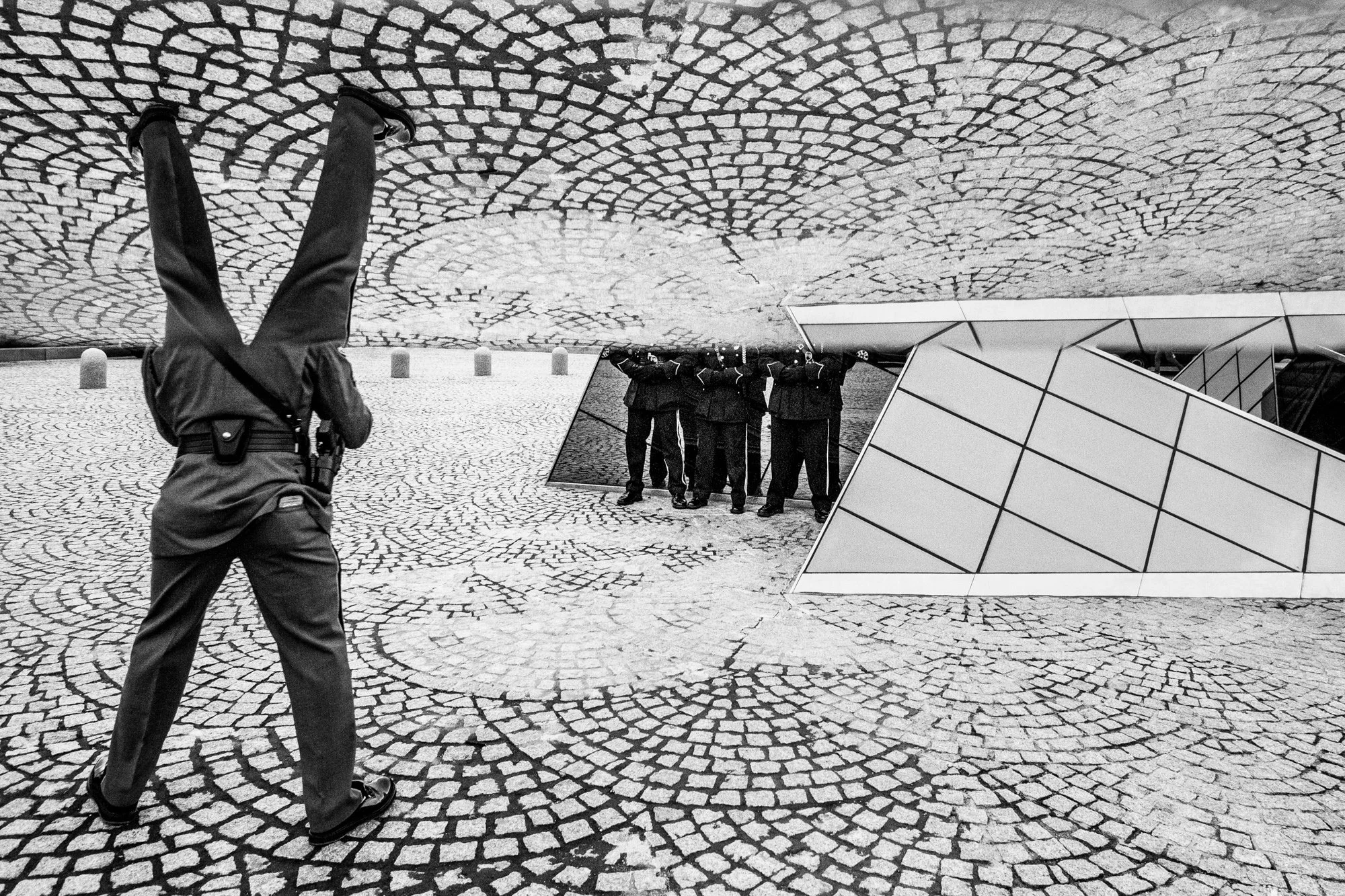 Black and white photo of a person walking in an outdoor urban area with patterned cobblestone pavement, modern building with geometric panels, and a reflective surface showing a group of people standing together.