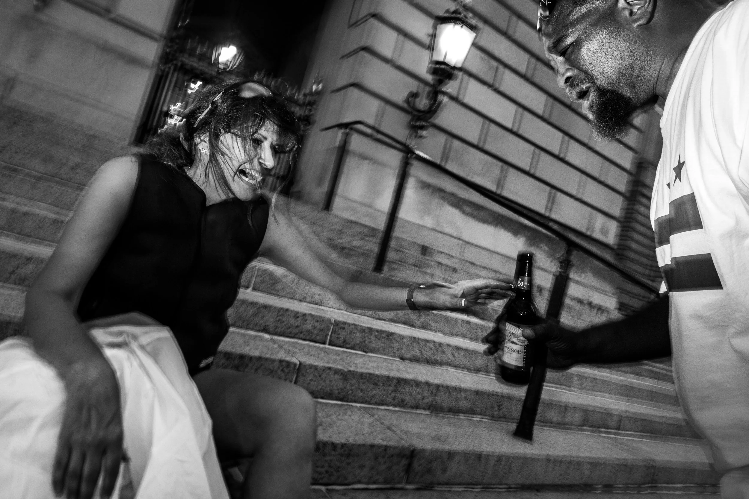 A woman sitting on stairs, reaching out to a man who is holding a beer bottle. The woman appears to be upset, and the man is looking down. The scene is in black and white, and there are lamps mounted on the wall behind them.