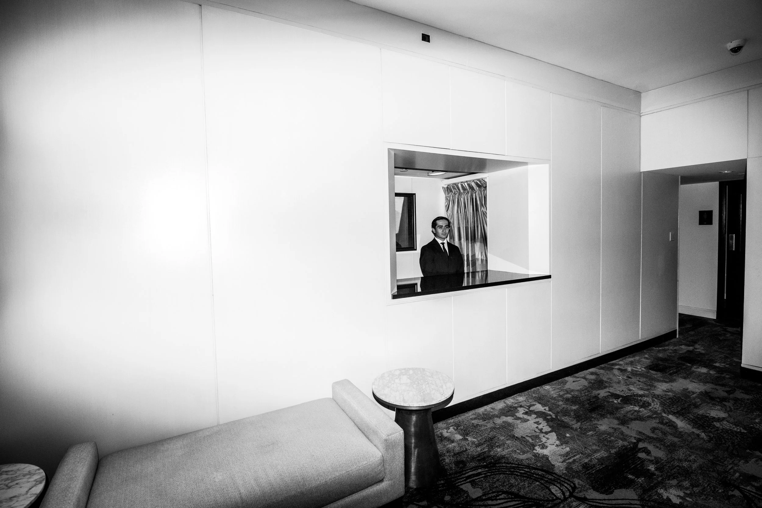 Black and white image of an indoor space featuring a reception desk with a man standing behind it, beige couches, small round marble-top tables, and a patterned carpet.
