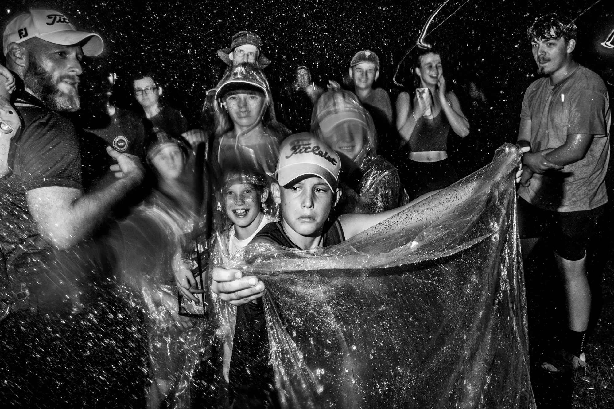Group of children and adults wearing rain gear and hats, standing together in a pouring rain, holding a clear plastic sheet, with water splashing around them.