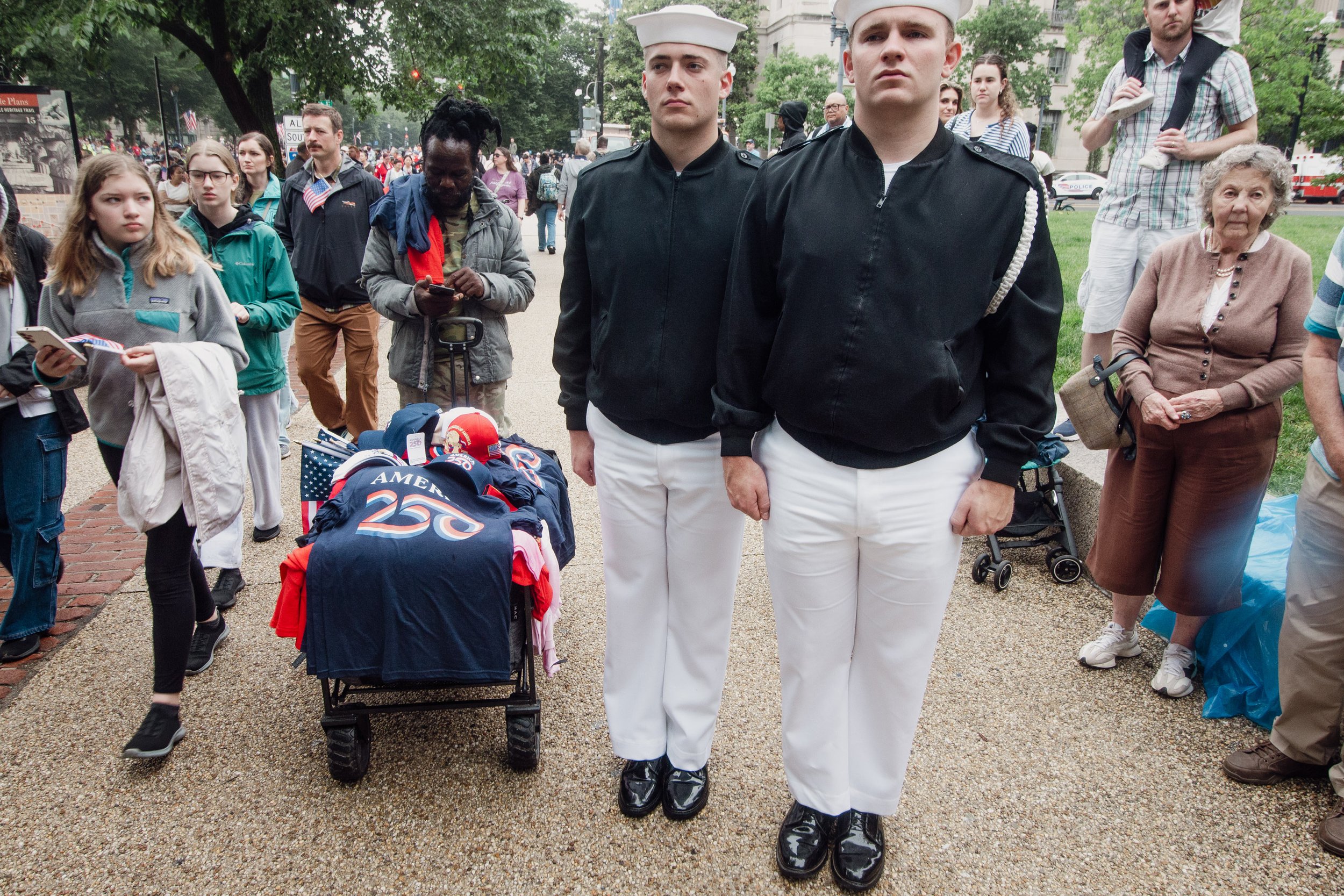 Young people, including two men in naval uniforms, standing among a crowd at a public event with trees and a building in the background.