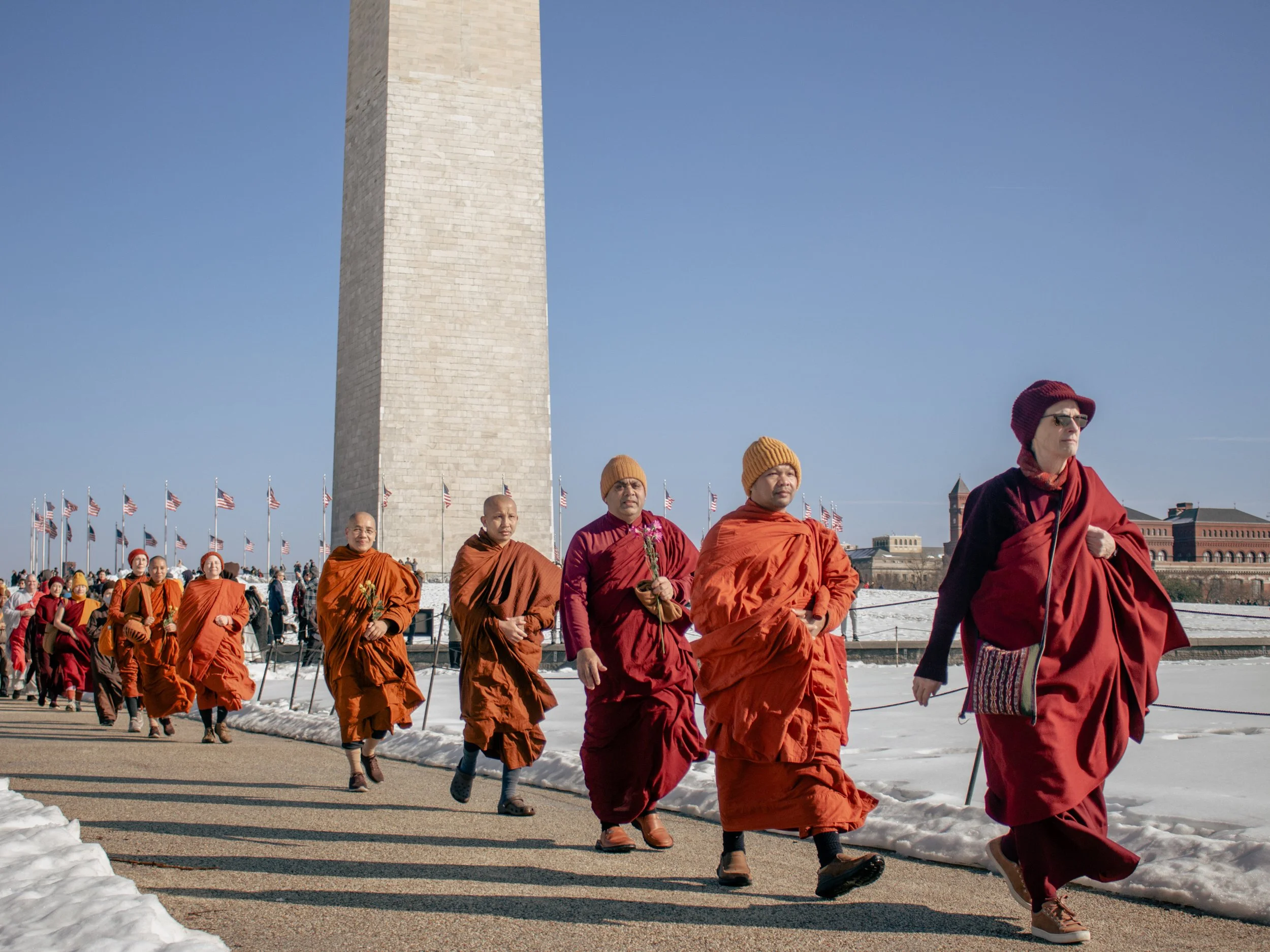 A line of Buddhist monks in orange and maroon robes walking outdoors on a clear day near the Washington Monument, with American flags in the background and snow on the ground.