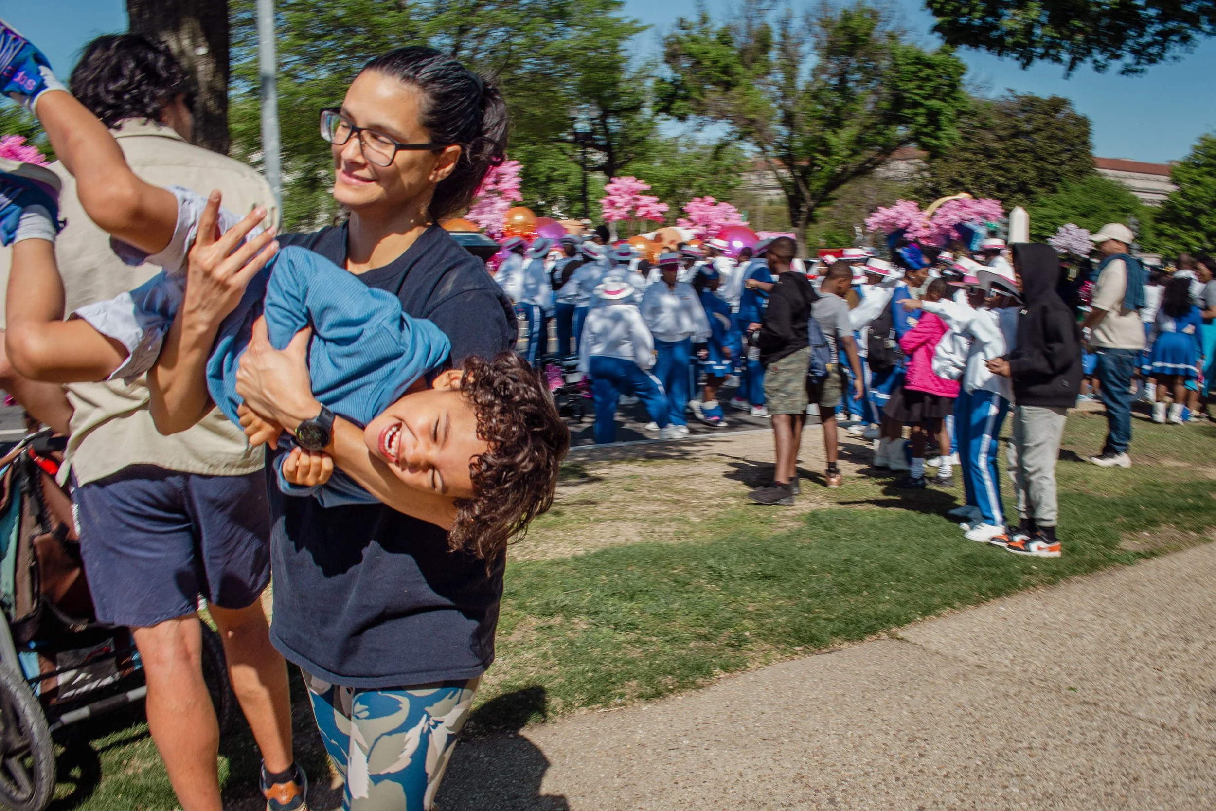 A woman smiling and wearing glasses holds a young boy with curly brown hair, who is smiling and leaning back, at an outdoor event with a crowd and pink flowers in the background.