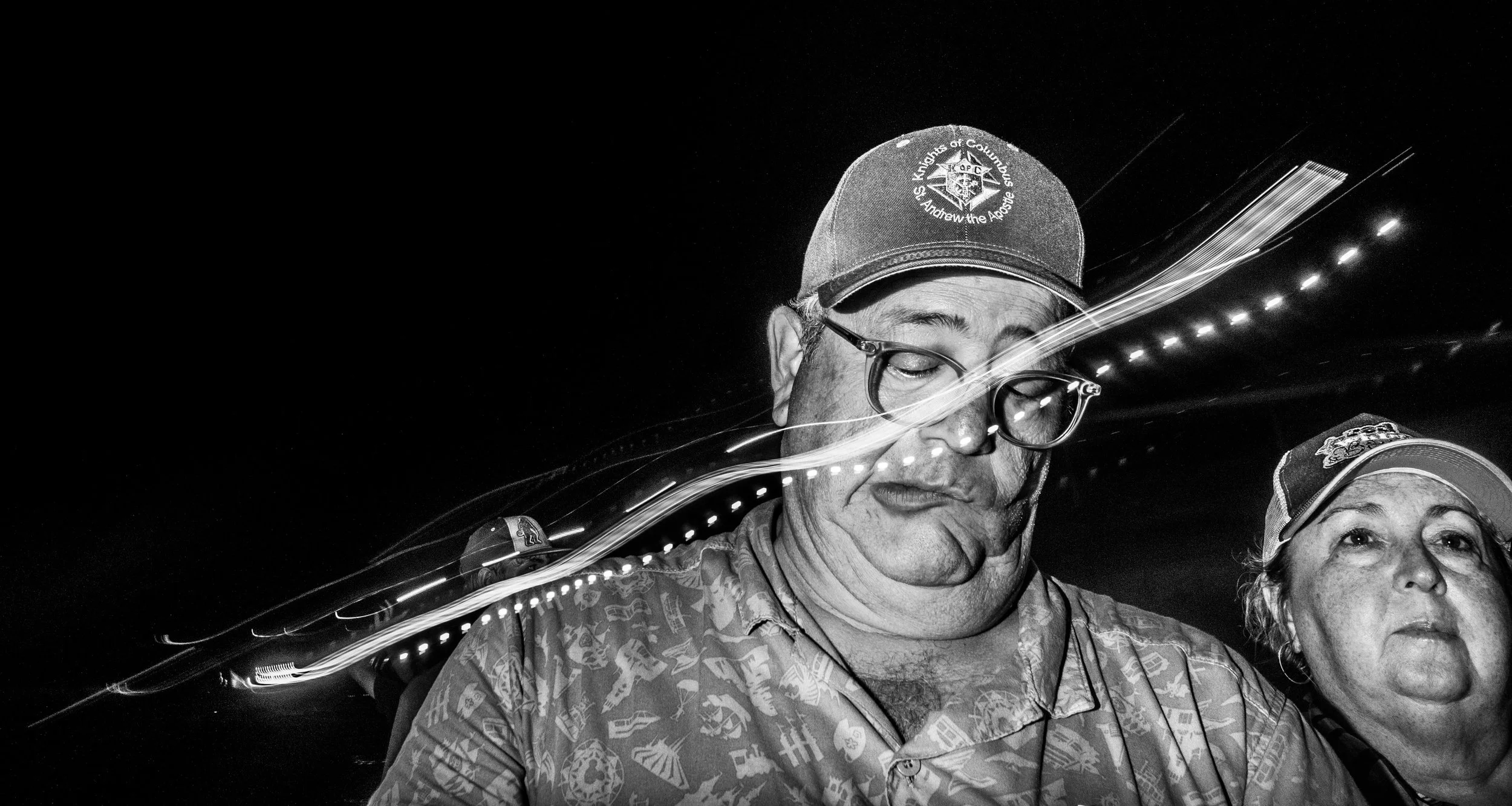 A black and white photo of two people, a man and a woman, at night. The man is wearing a cap with a Columbus, Ohio, police emblem, glasses, and has his eyes closed. The woman beside him is also wearing a cap and has her eyes open.