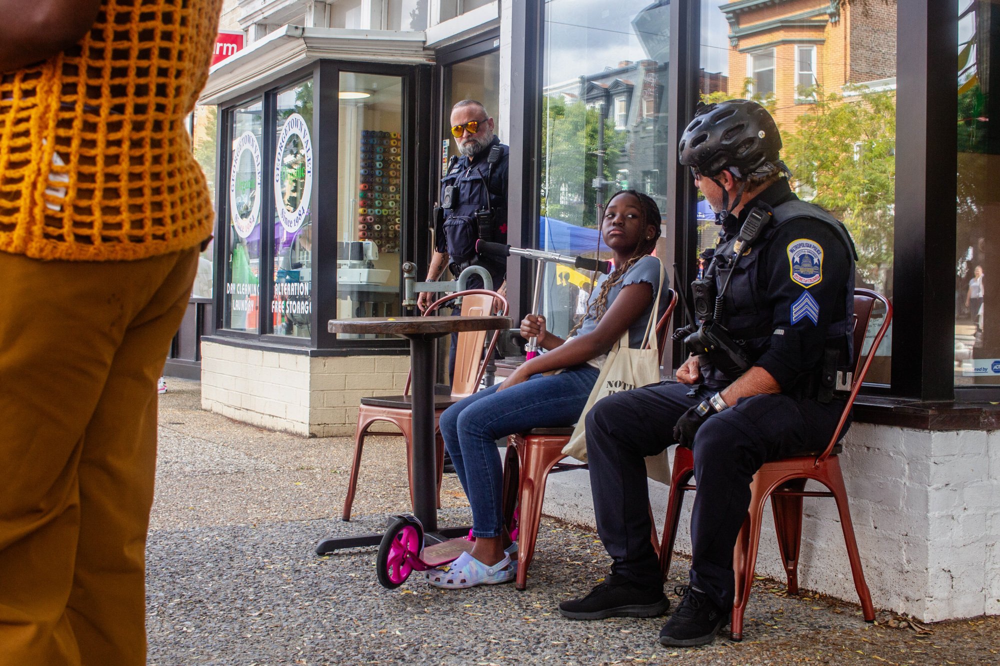 Two police officers, a woman and a man, sitting outside a storefront with glass windows, talking to a girl sitting on a chair. The girl has braided hair, is wearing a gray shirt and blue jeans, and has a pink scooter at her feet. The storefront has signs advertising laundry and storage services, and there are trees and buildings reflected in the glass.