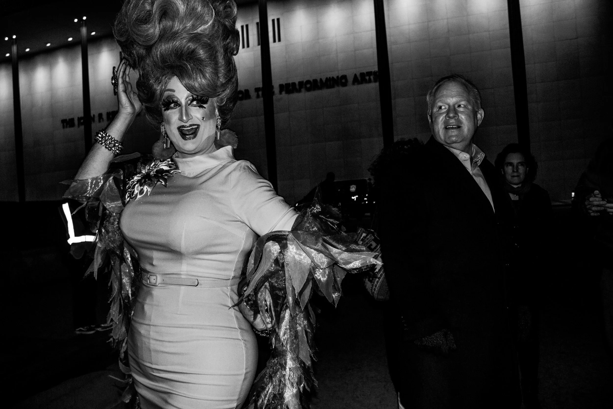 A drag queen with elaborate hair and makeup, wearing a shiny dress and ruffled sleeves, smiling and looking to the side at an event or performance, with a man in a suit and a person in the background.
