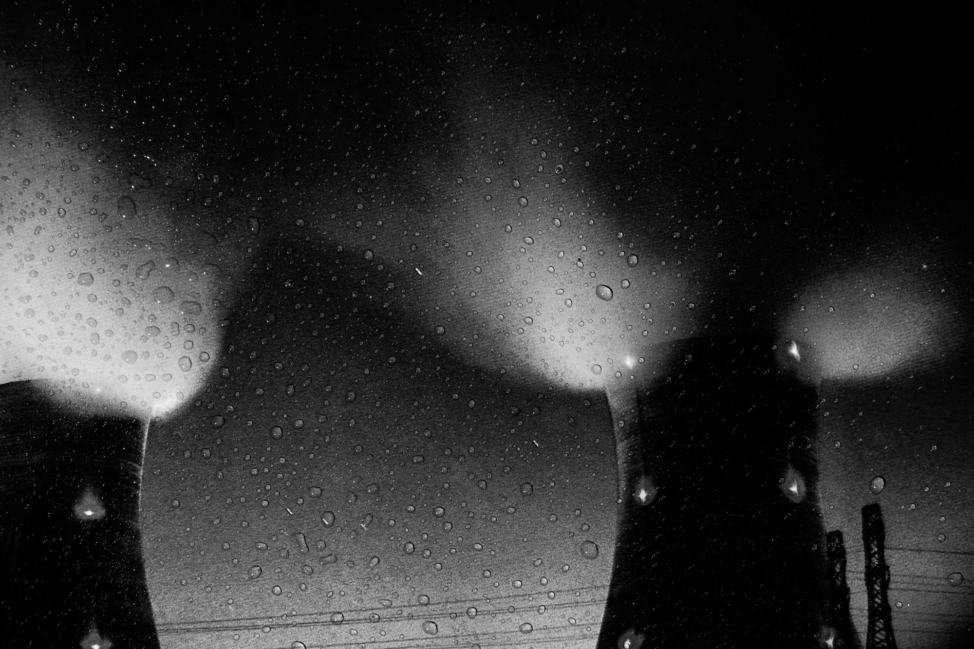 Close-up of a water-covered window with visible raindrops on the glass and some blurry nighttime city lights and streetlights in the background.