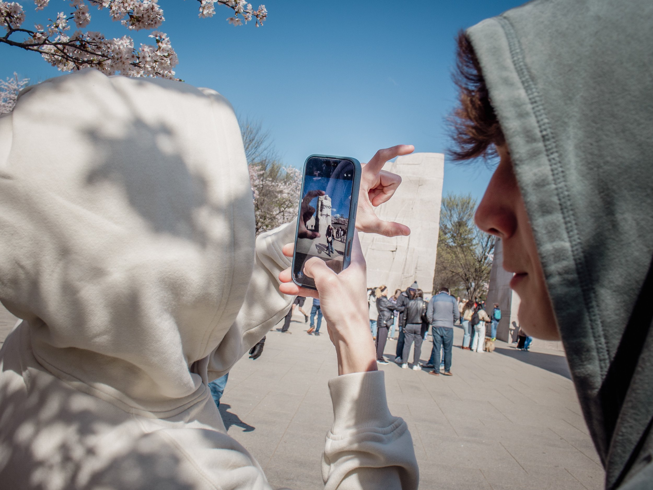 A person wearing a cream-colored hoodie is taking a photo of the Washington Monument with a smartphone. The photo captures the monument and pink blossoming trees in the background, with other visitors walking around on a sunny day.