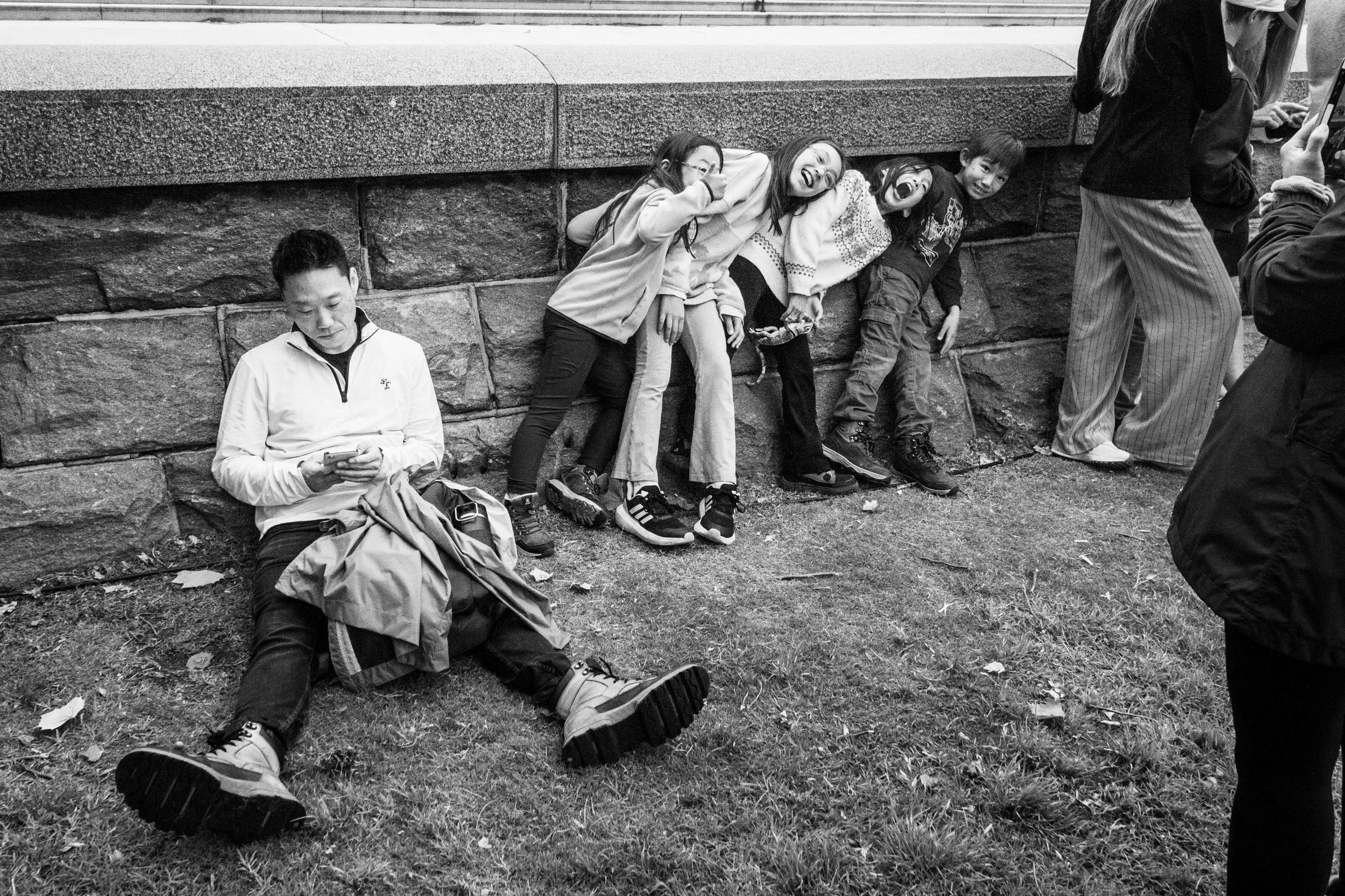 A group of five people sitting and standing against a stone wall in a park, some smiling and laughing, while others are looking at their phones.