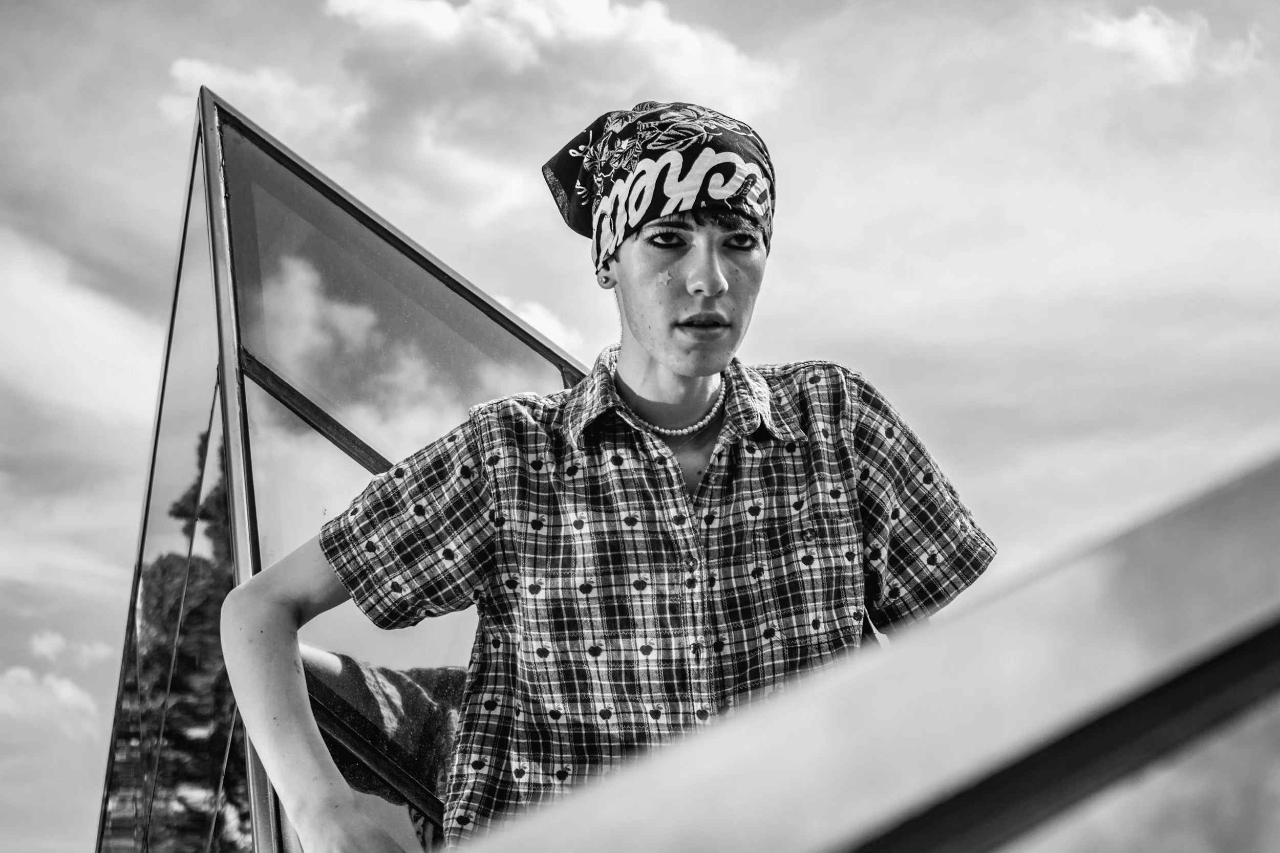 Black and white photo of a young person wearing a checked shirt, a headscarf, and a pearl necklace, standing outdoors with cloudy skies and trees in the background, posing with one hand on their hip and looking at the camera.