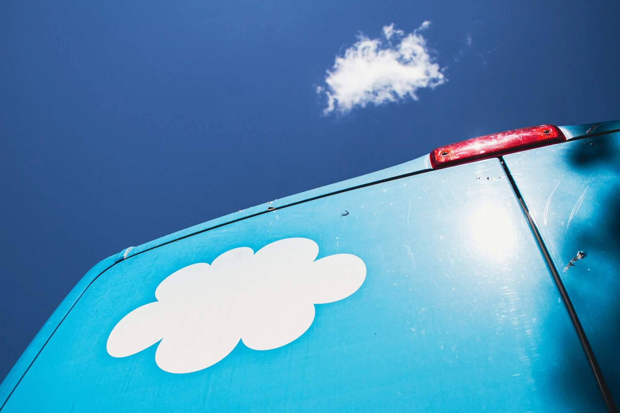 Close-up of the rear of a blue vehicle featuring a white cloud graphic, with a bright red tail light and a clear blue sky with some clouds in the background.