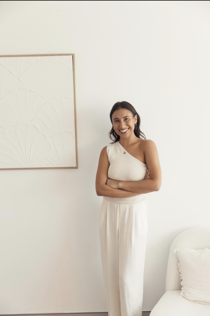 Woman standing against a white wall, smiling with arms crossed, dressed in a white one-shoulder top and matching wide-leg pants, next to a framed abstract artwork and a white sofa with a pillow.