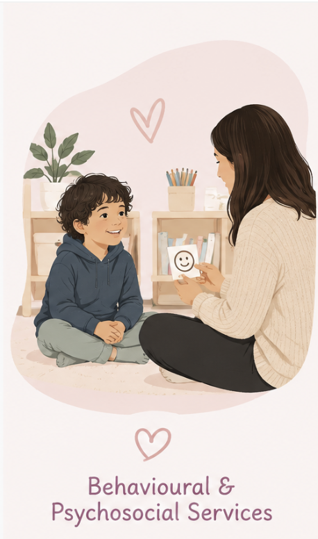 A woman and a young boy sitting on the floor, engaging in a therapy session. The woman holds a card with a smiley face. The background features bookshelves, a potted plant, and a pink heart drawing on the wall. The text reads "Behavioral & Psychosocial Services."