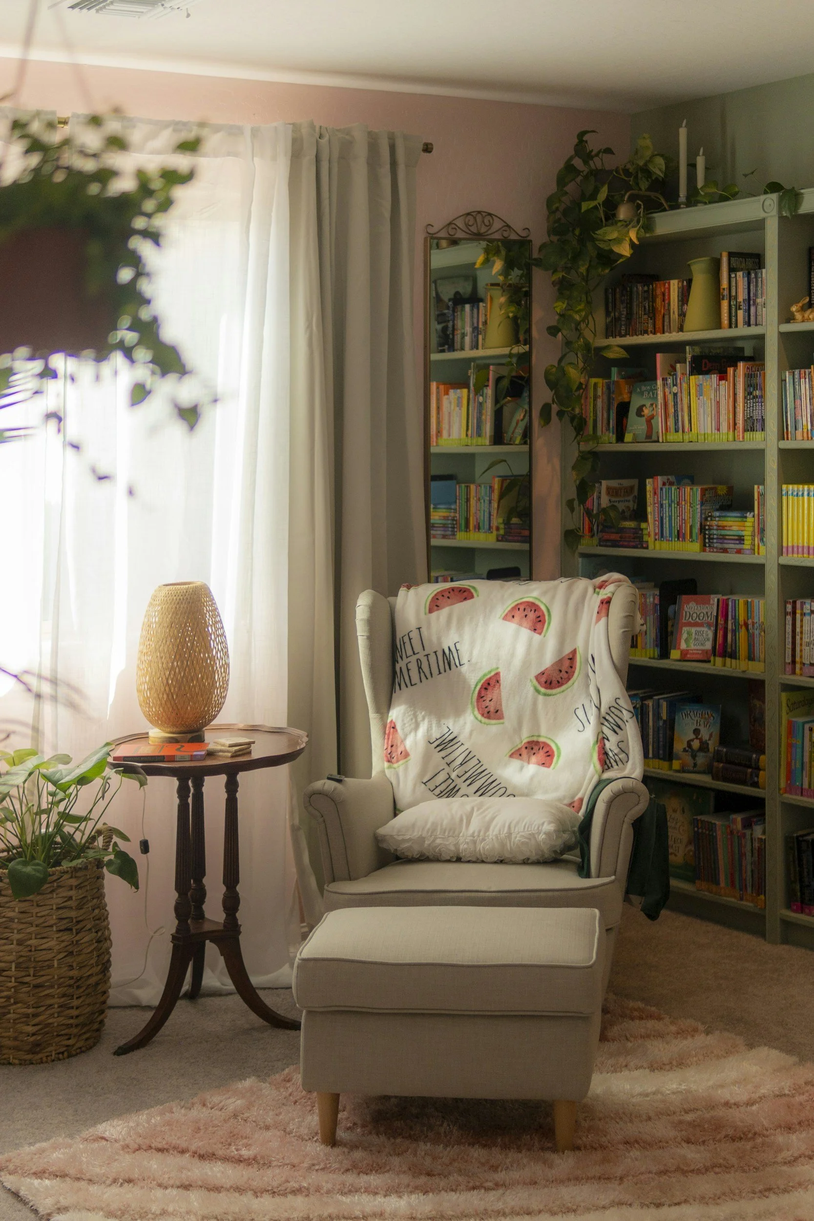 A cozy reading nook featuring a white armchair with a watermelon-themed throw blanket and matching pillow, a small wooden side table with a woven table lamp, a bookshelf filled with books, and a pink fluffy rug on a beige carpeted floor. Sunlight filters through white curtains covering a window.