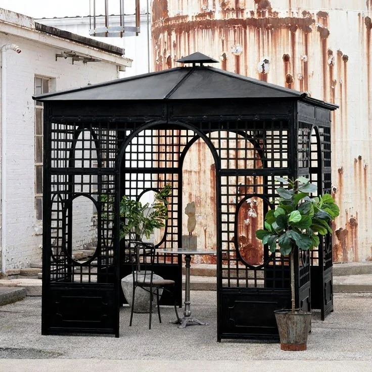 A black metal gazebo with decorative lattice walls, set outdoors on a paved surface. Inside, there is a small round table with a decorative sculpture and a chair, along with potted green plants nearby. In the background, a rusted metal tank and a white brick building are visible.