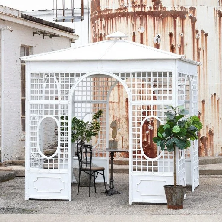 White outdoor gazebo with lattice sides in an urban setting, decorated with a potted plant and chairs.