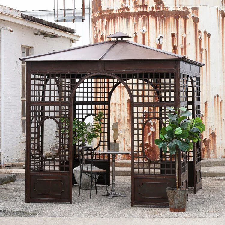 A small outdoor gazebo with a dark wooden lattice structure and a sloped roof, placed on a paved surface in front of a rusted metal tank. Inside the gazebo, there are two chairs, a small table, a stone sculpture, and a potted ficus plant outside the entrance.