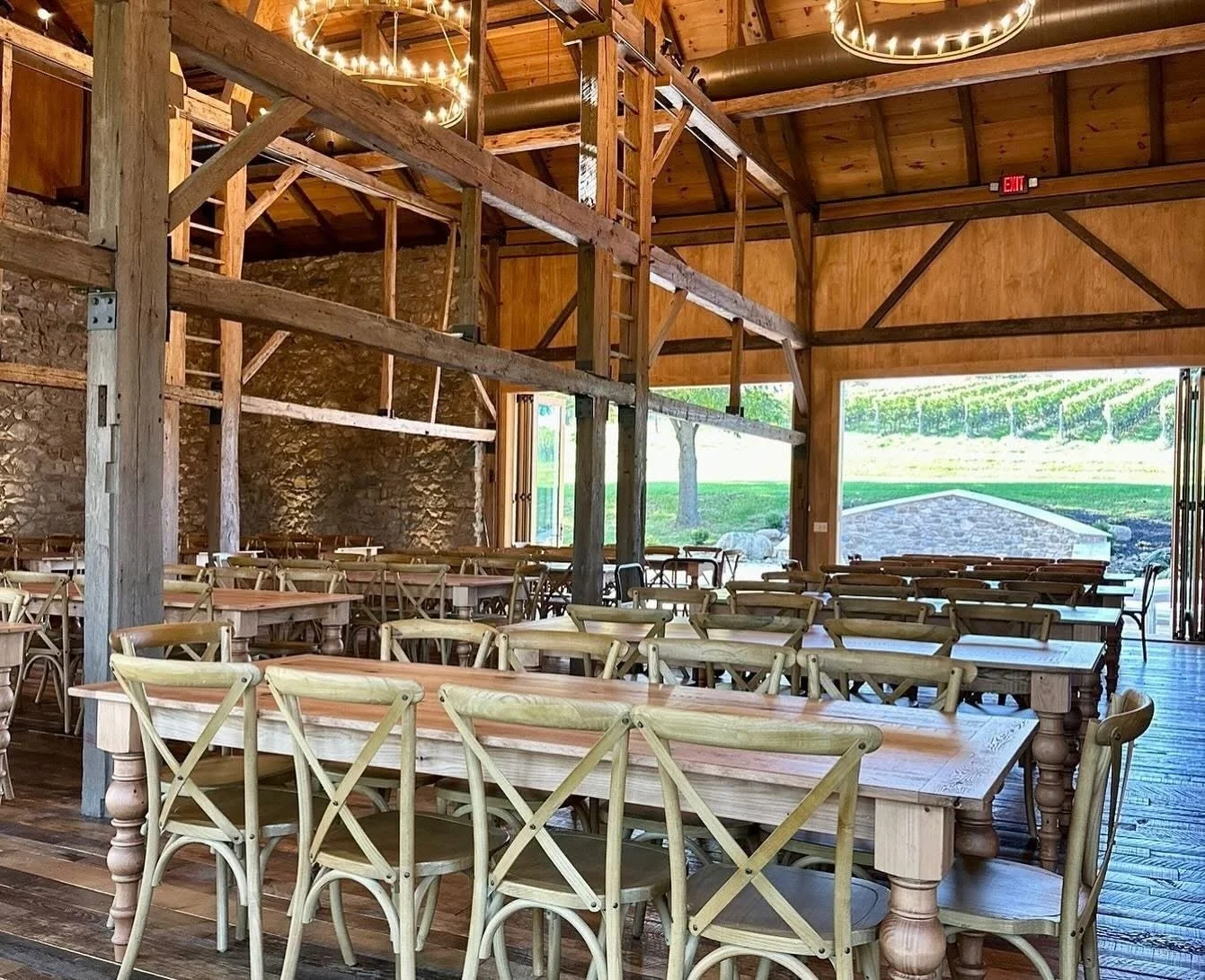 Interior of a rustic wooden event space with long tables and chairs, exposed stone wall, large open windows, and a view of green outdoor scenery.
