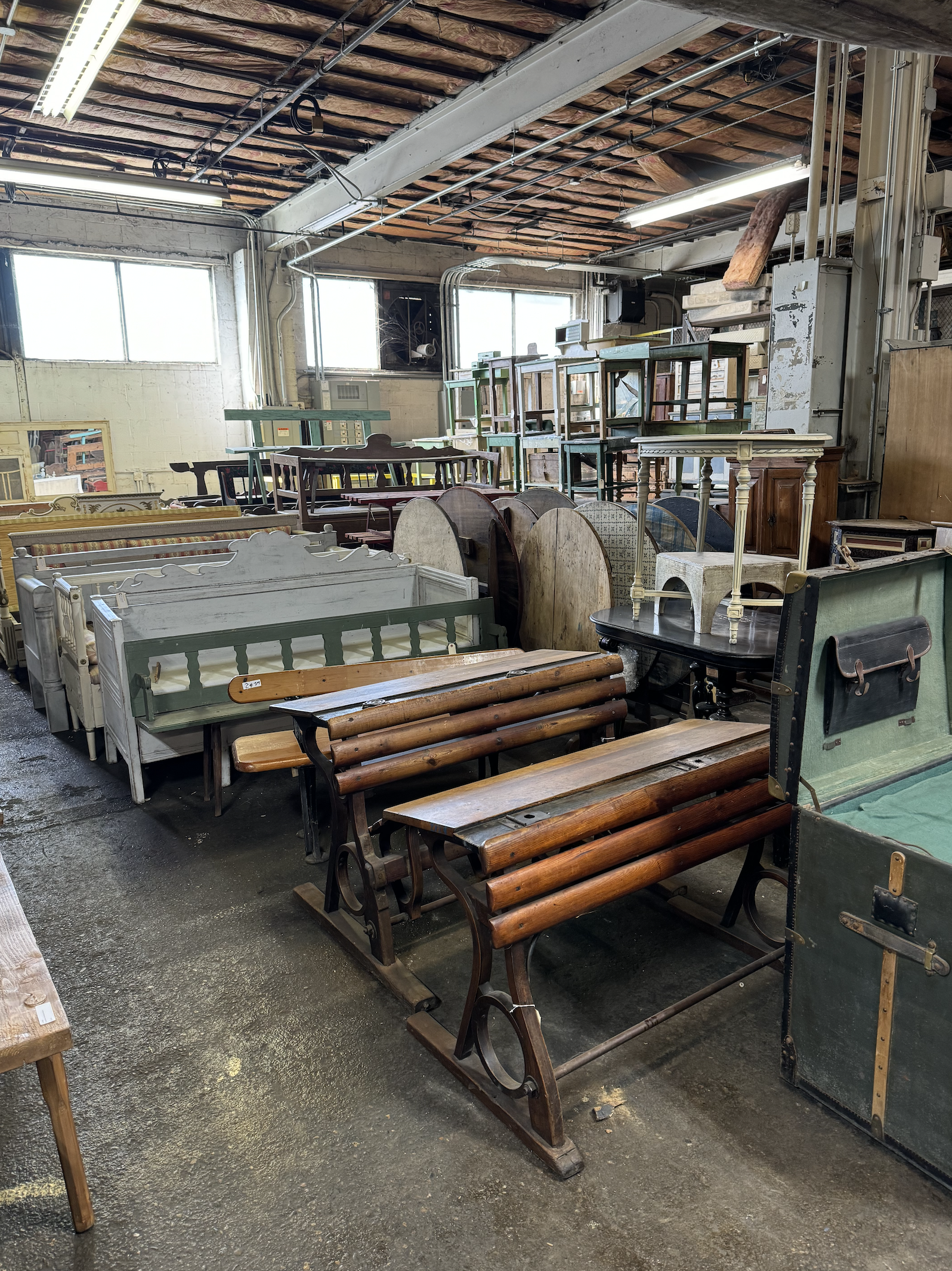 Various vintage and antique furniture pieces, including benches, tables, and chairs, are arranged in a warehouse or thrift store with exposed brick and wooden ceiling.
