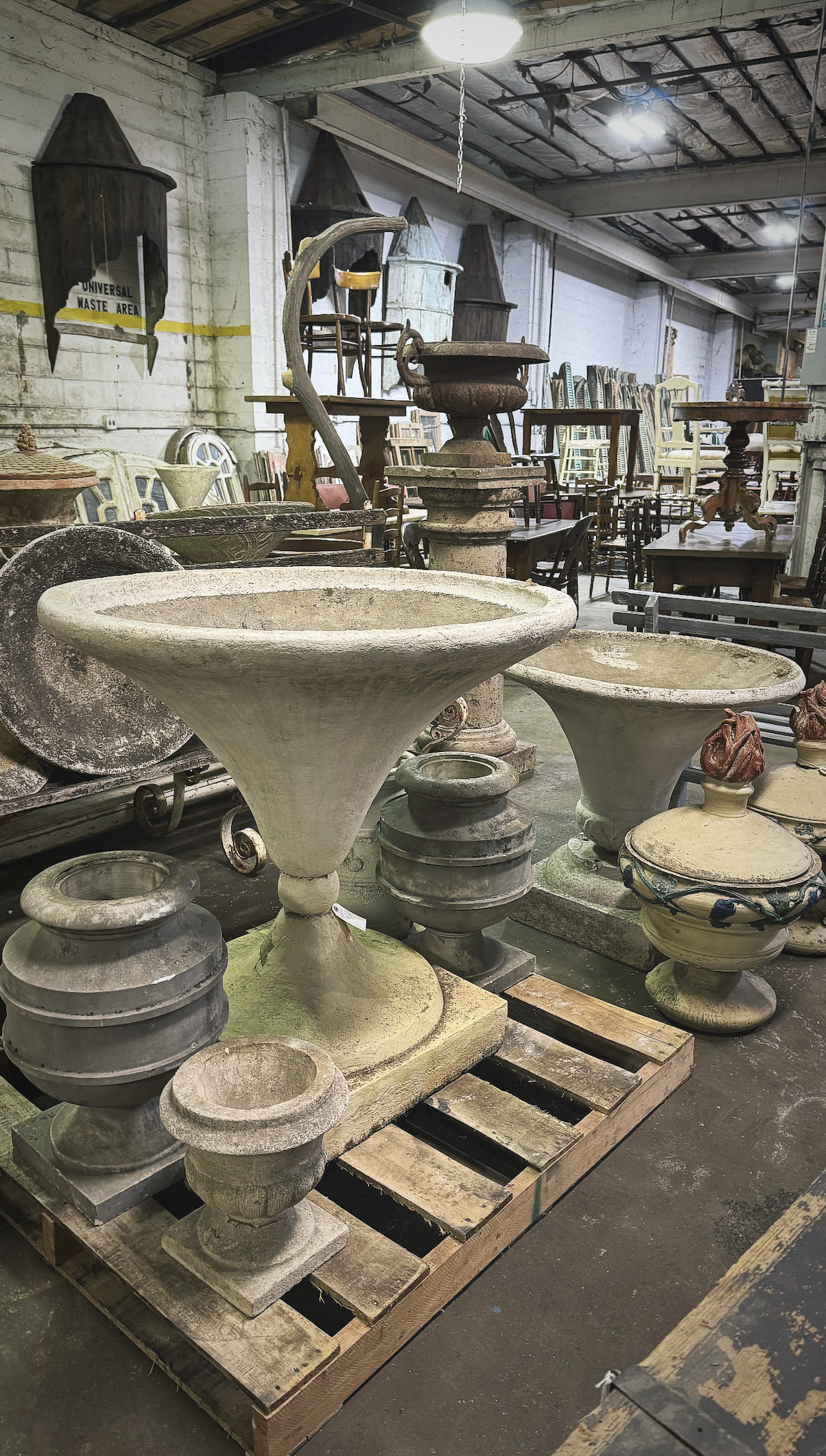 Indoor space filled with various antique and vintage planters and garden urns, some resting on wooden pallets and tables, with chairs and cabinets in the background, and a high ceiling with exposed beams.