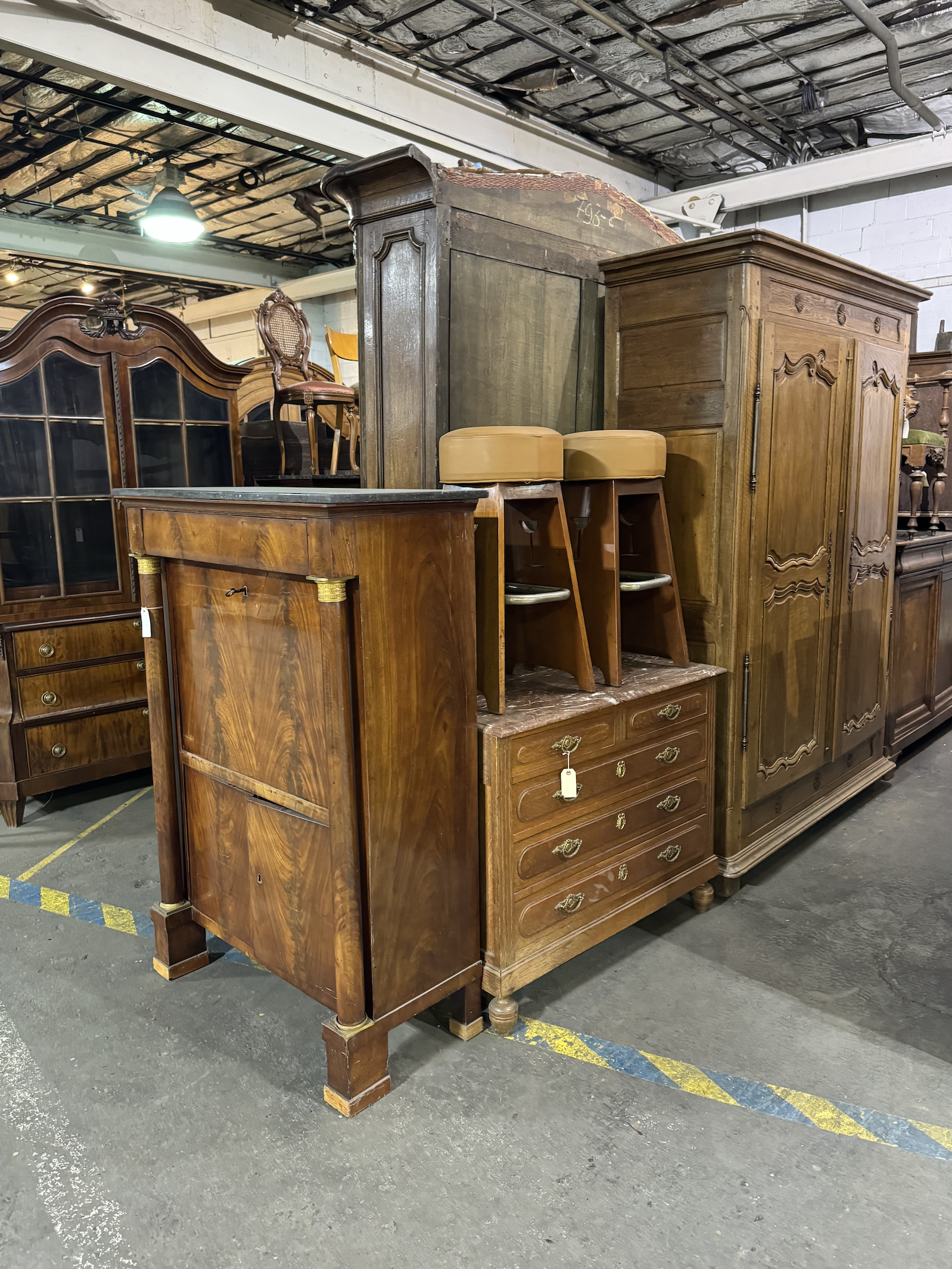 A collection of antique wooden furniture including dressers, cabinets, and chairs stacked in a warehouse.