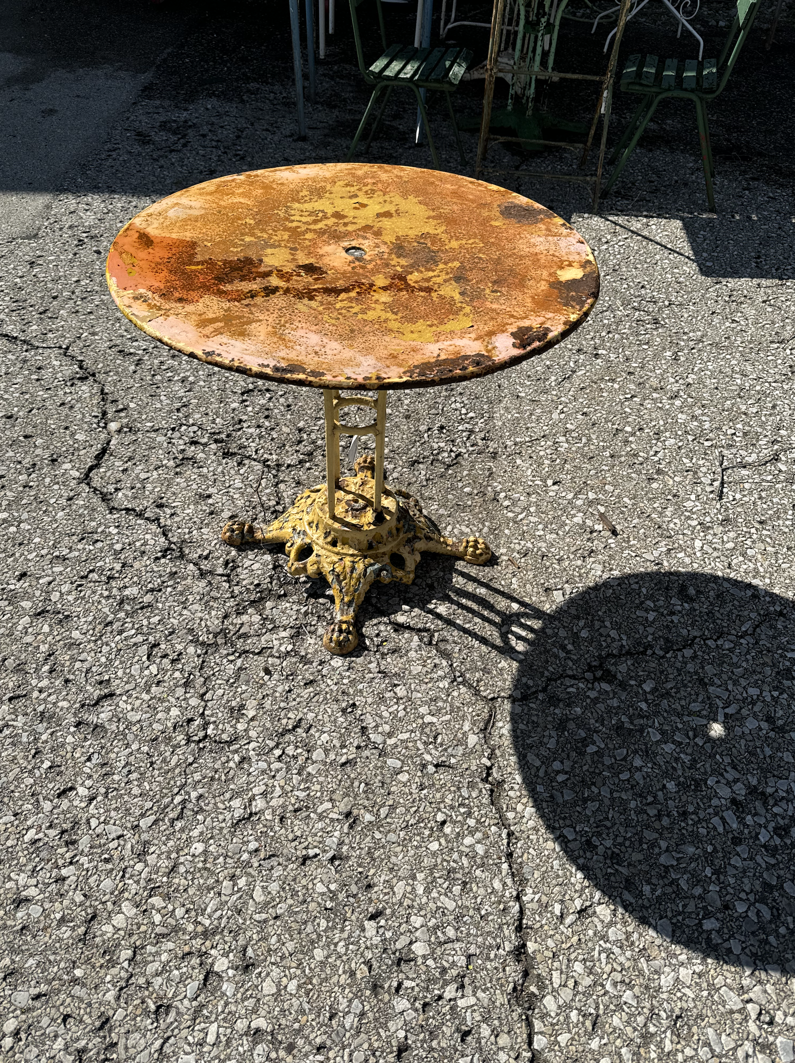 A round metal table with a rusted orange and yellow surface, situated on cracked asphalt. The table has a decorative yellow base and casts a shadow in the sunlight.