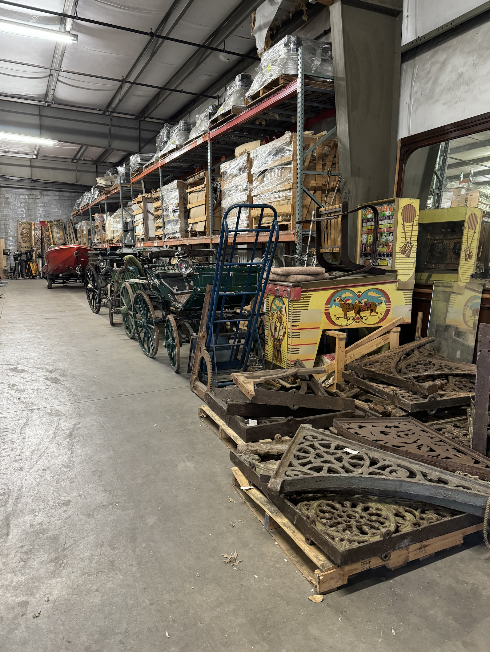 Interior of a warehouse storage area with vintage ride-on toys, including a small horse-drawn car and other antique amusement rides, along with metal and wooden decorative panels on pallets.