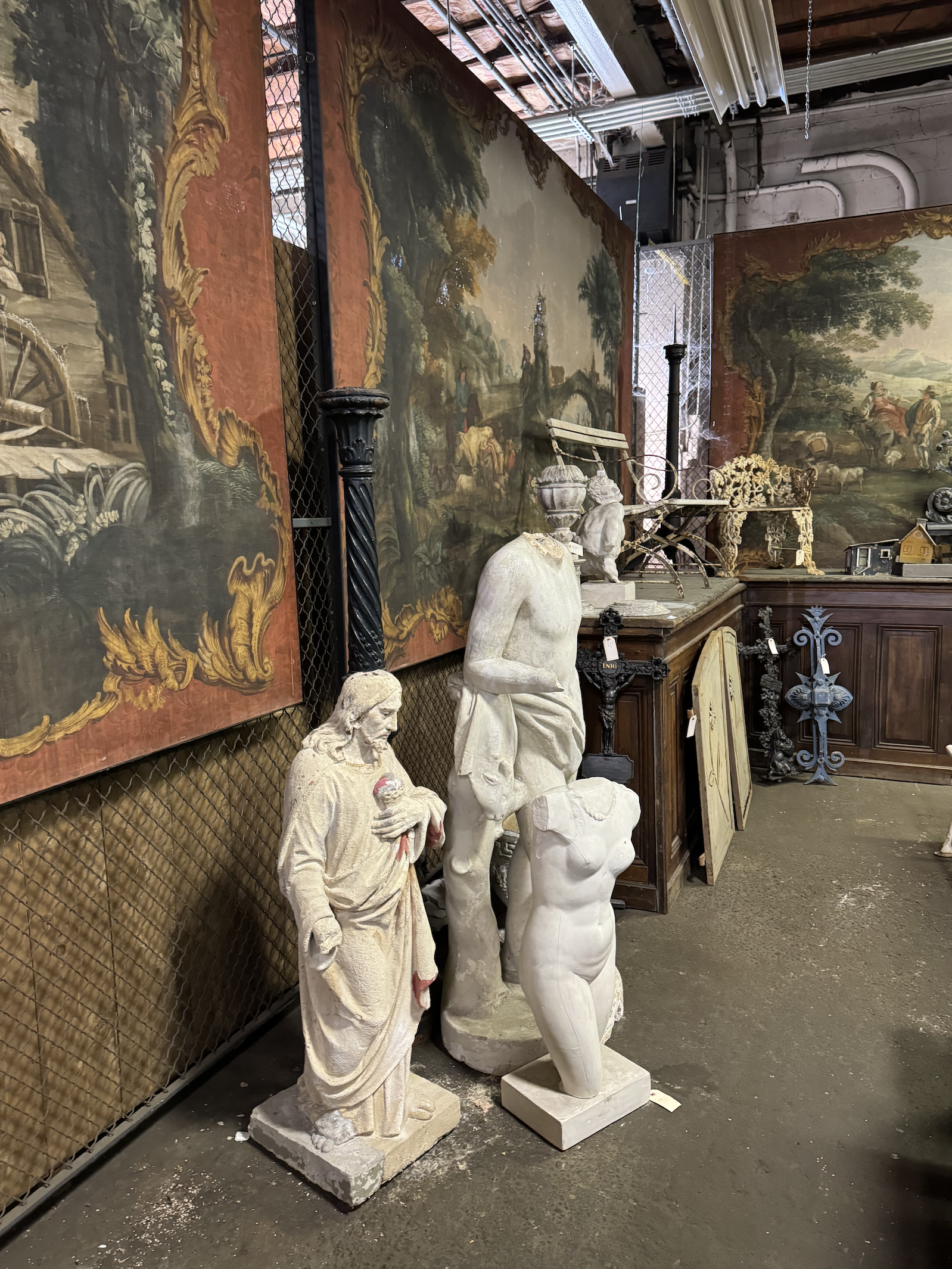 Sculptures of Jesus holding a heart, a headless female torso, and a headless male torso in an antique store surrounded by vintage paintings and furniture.