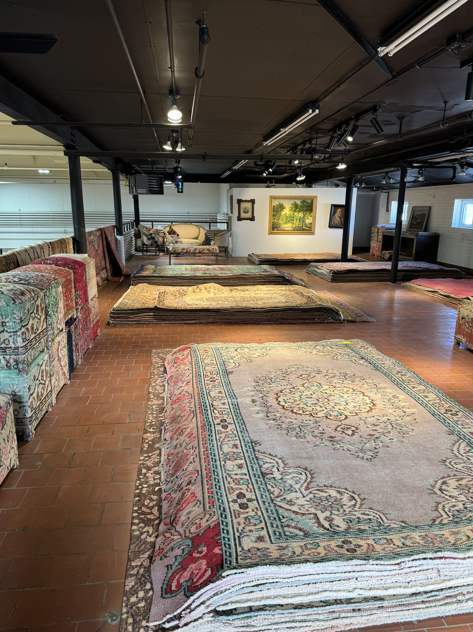A room with many stacked and rolled antique rugs and carpets, along with some furniture and framed paintings on the walls.
