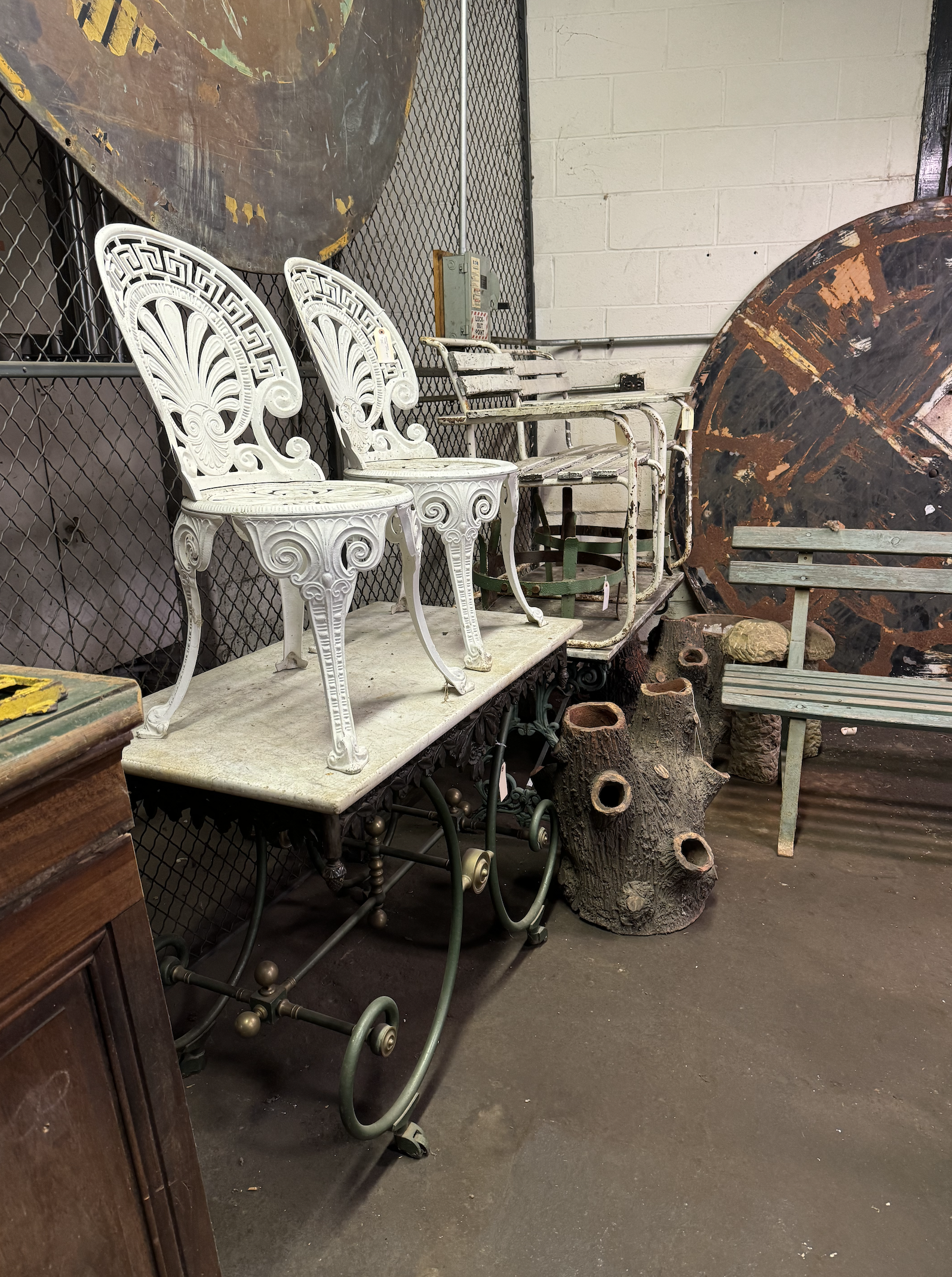 A collection of vintage furniture including two ornate white chairs, a worn green bench, and a wooden log with multiple holes, all set against a black metal fence and white brick wall in a warehouse or thrift store.