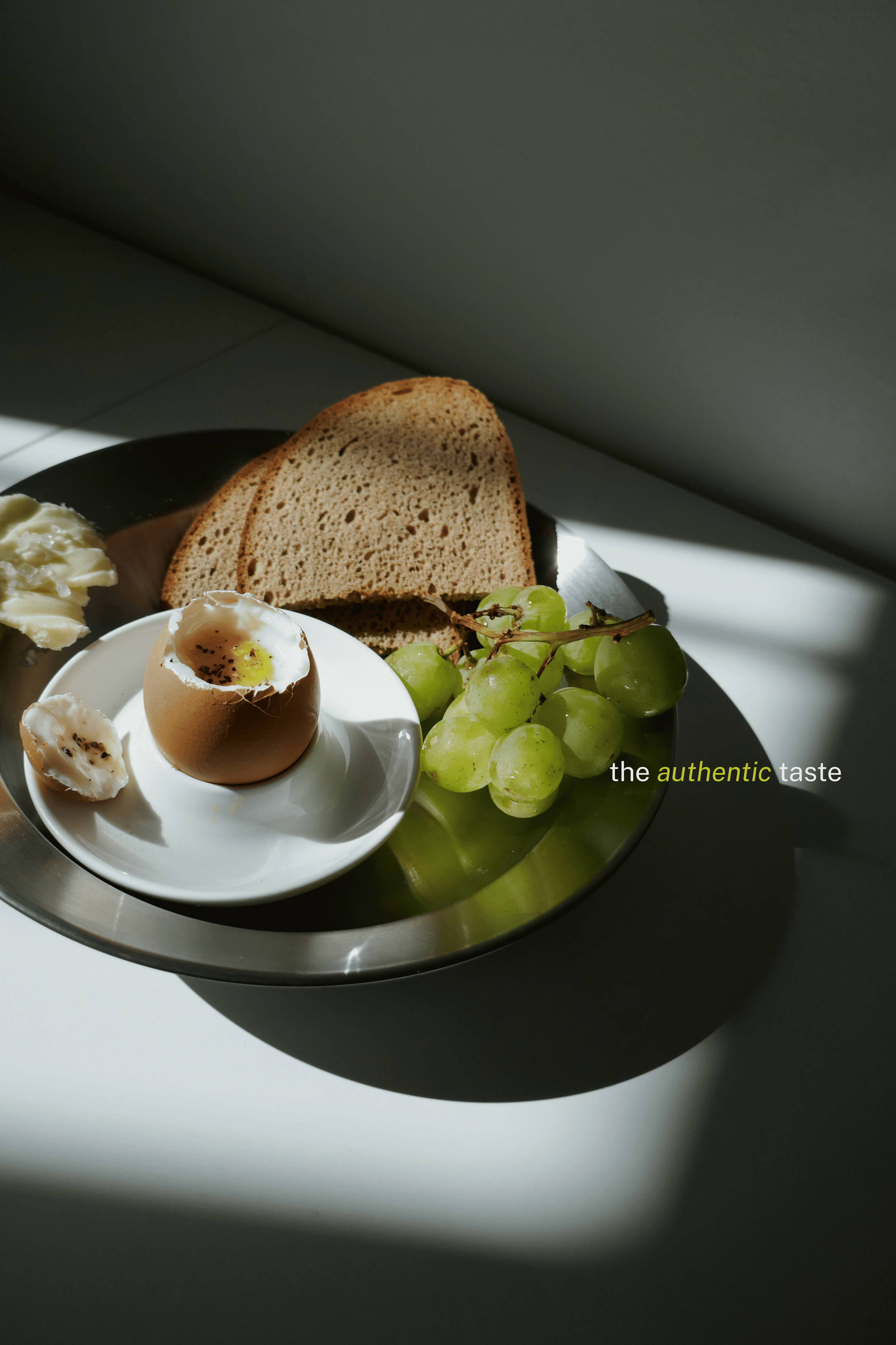 Uovo sodo in una ciotola bianca, pane integrale, grappolo di uva verde, burro, e pane tostato su un piatto nero con sfondo grigio scuro e luce naturale