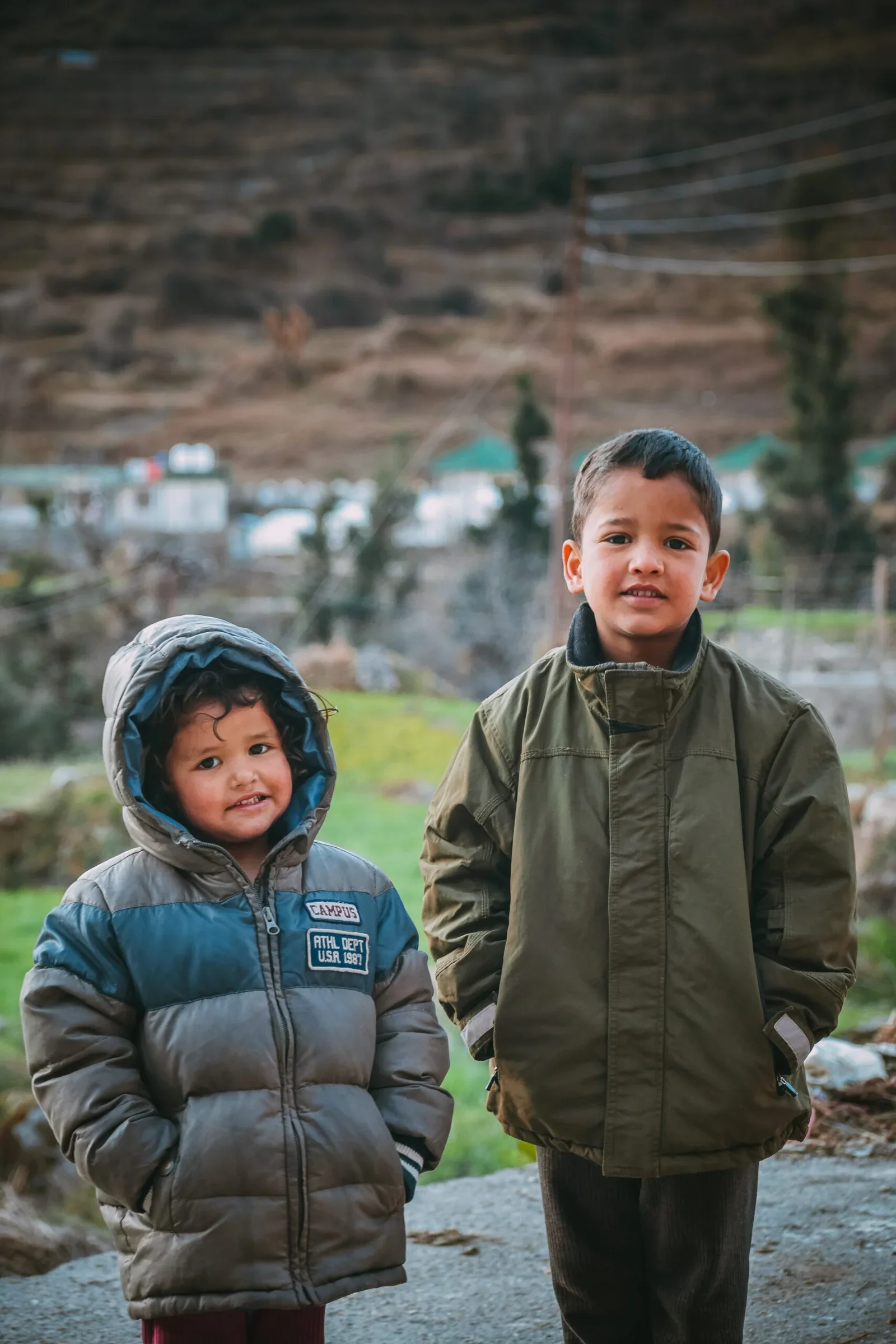 Two young boys standing outdoors during daytime, wearing jackets, with a mountainous landscape in the background.
