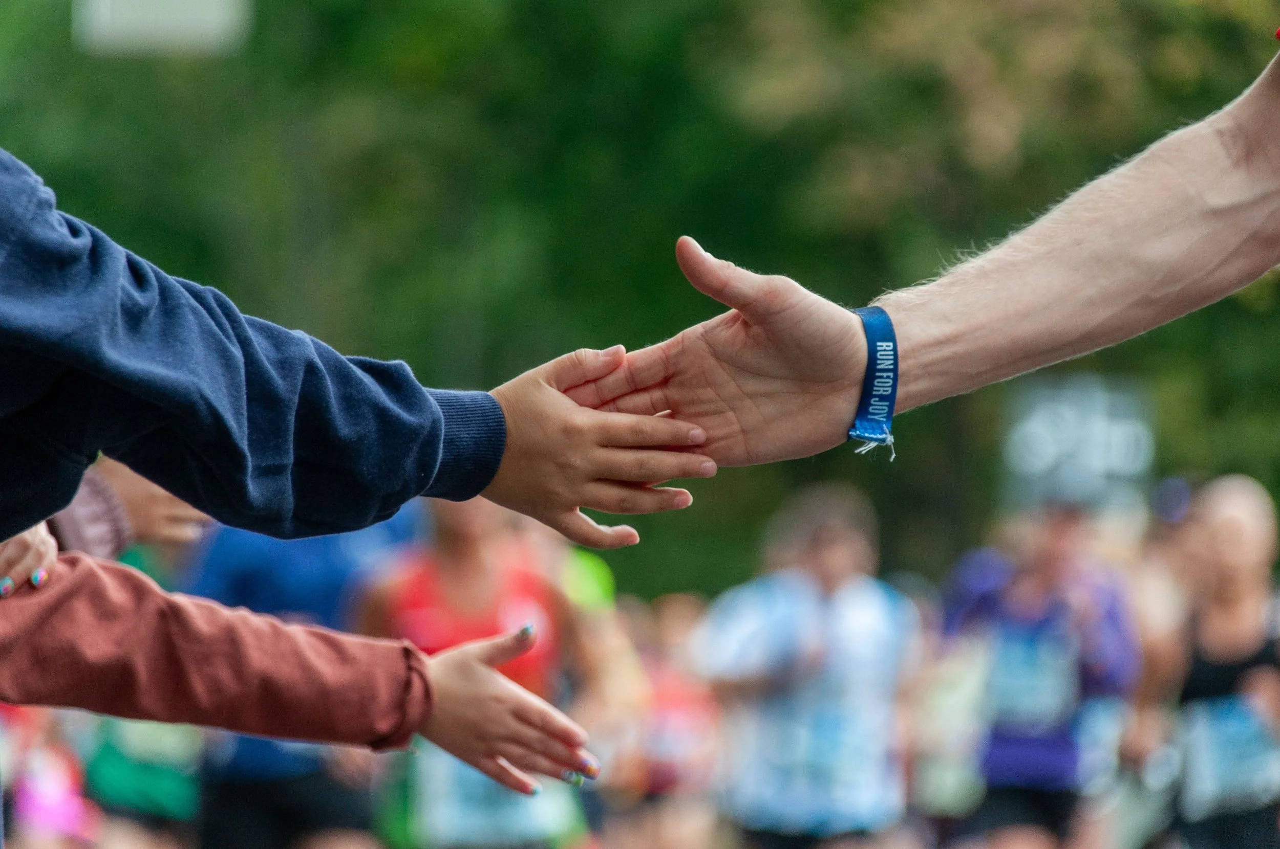 Close-up of children's hands reaching out for each other, with blurred children and greenery in the background.