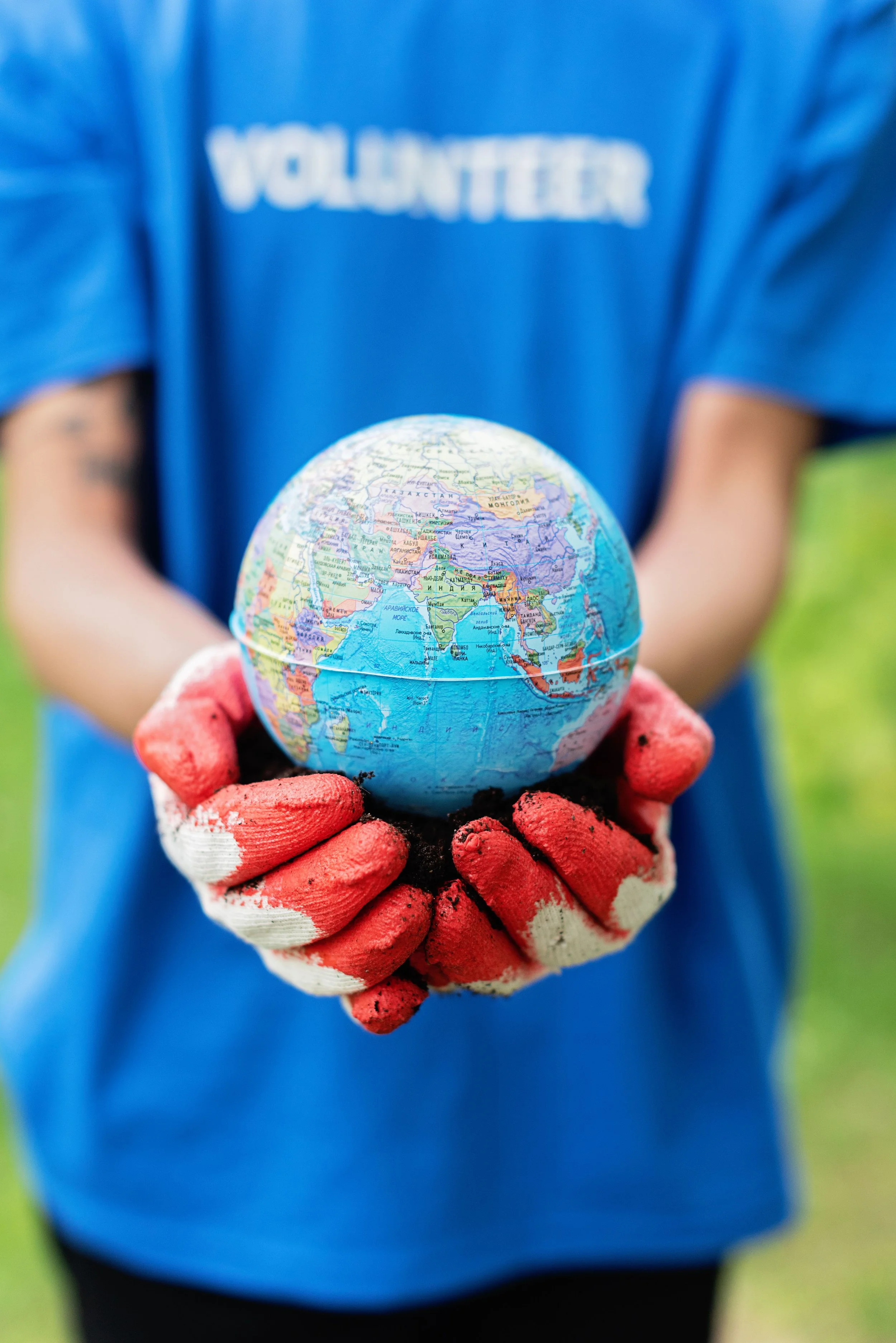 Person wearing a blue shirt with 'VOLUNTEER' written on it, holding a small globe or world map model with red gloves, outdoors with green grass in the background.