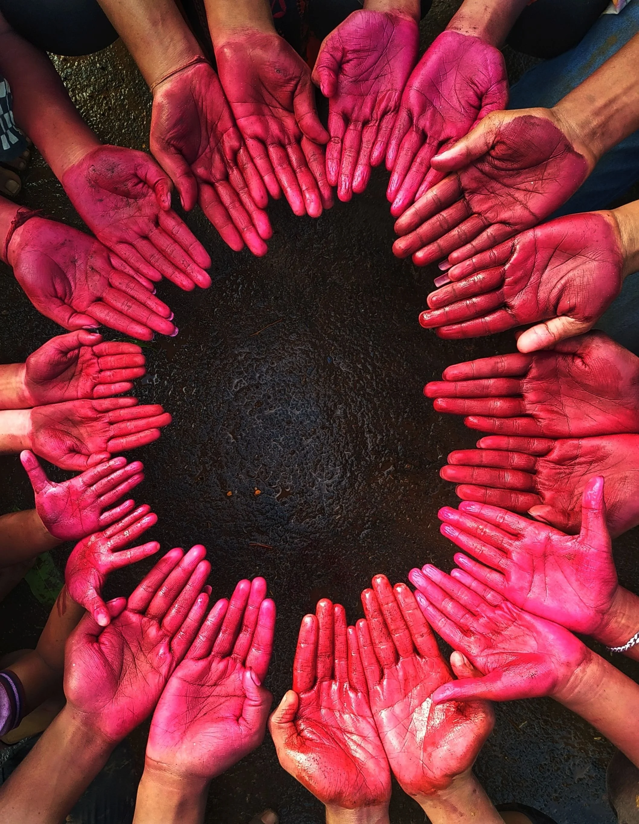 Multiple hands covered in pink and red pigment, arranged in a circle with palms facing up, on a dark surface.