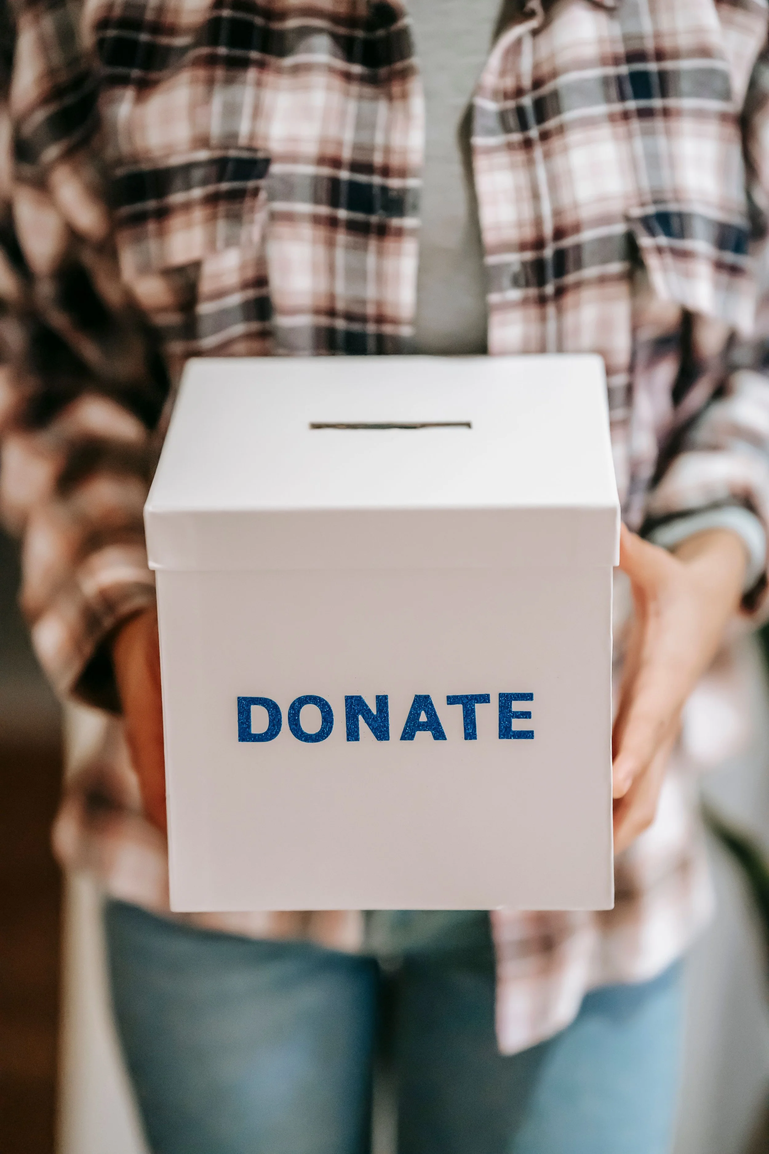 Person holding a white donation box with blue text that says 'DONATE', wearing a plaid shirt and jeans.