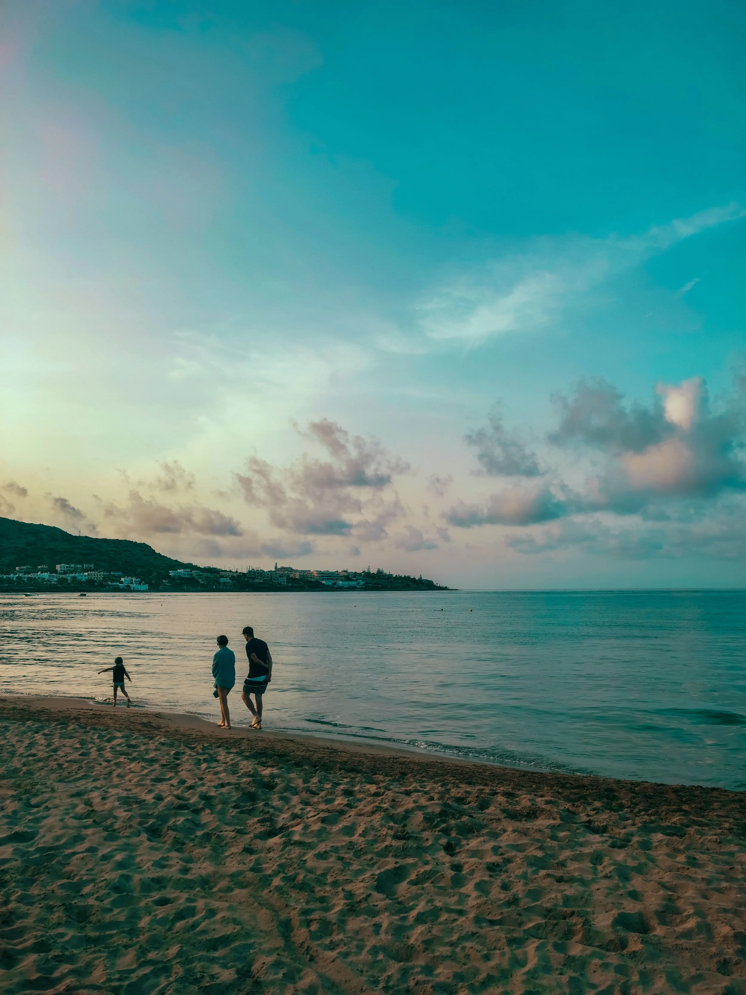 People walking along a sandy beach during sunset with calm water, small clouds, and a distant hillside with houses.