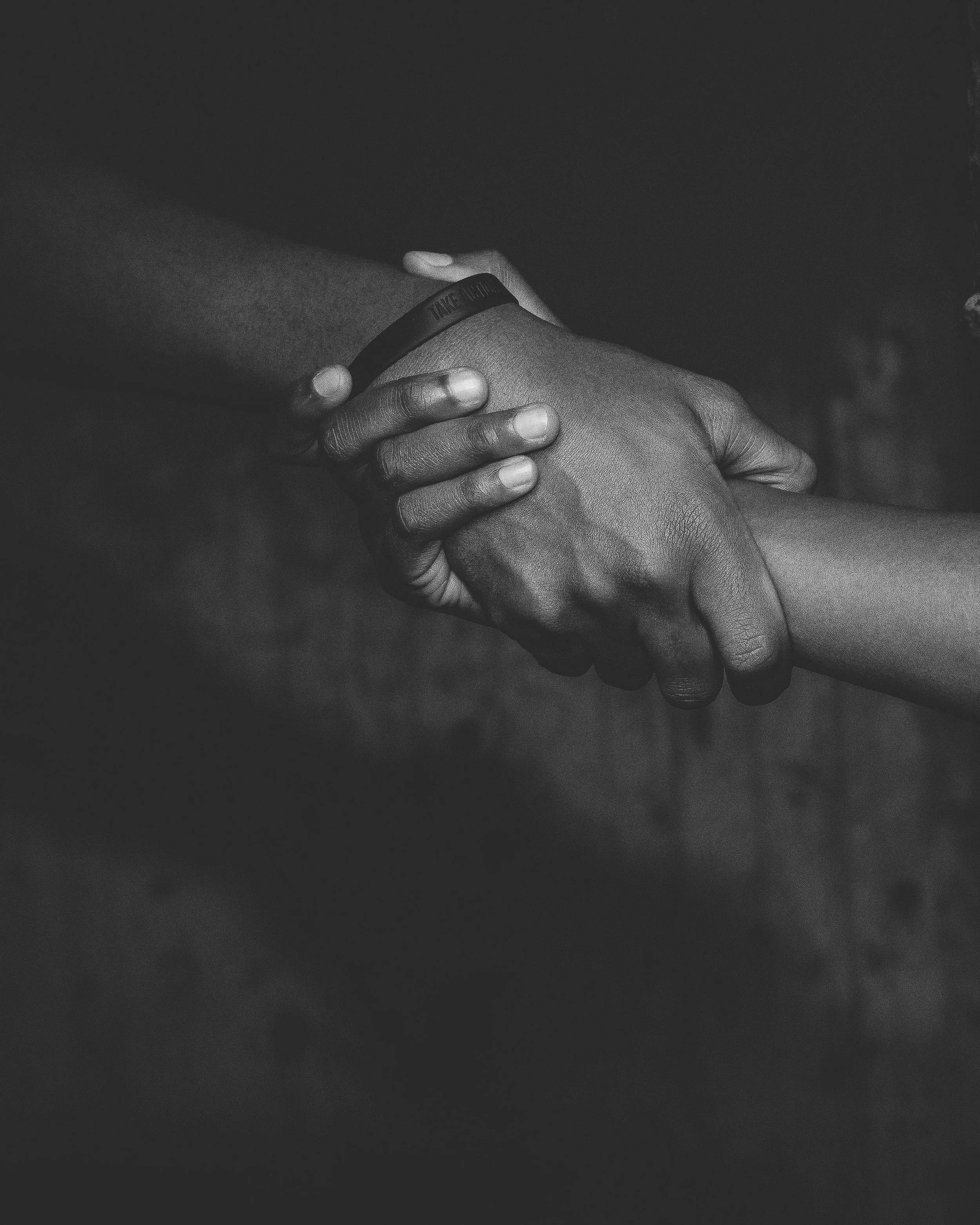 A black and white close-up photo of two hands clasped together, one hand wearing a wristband with the partially visible text 'TAKE MI...'