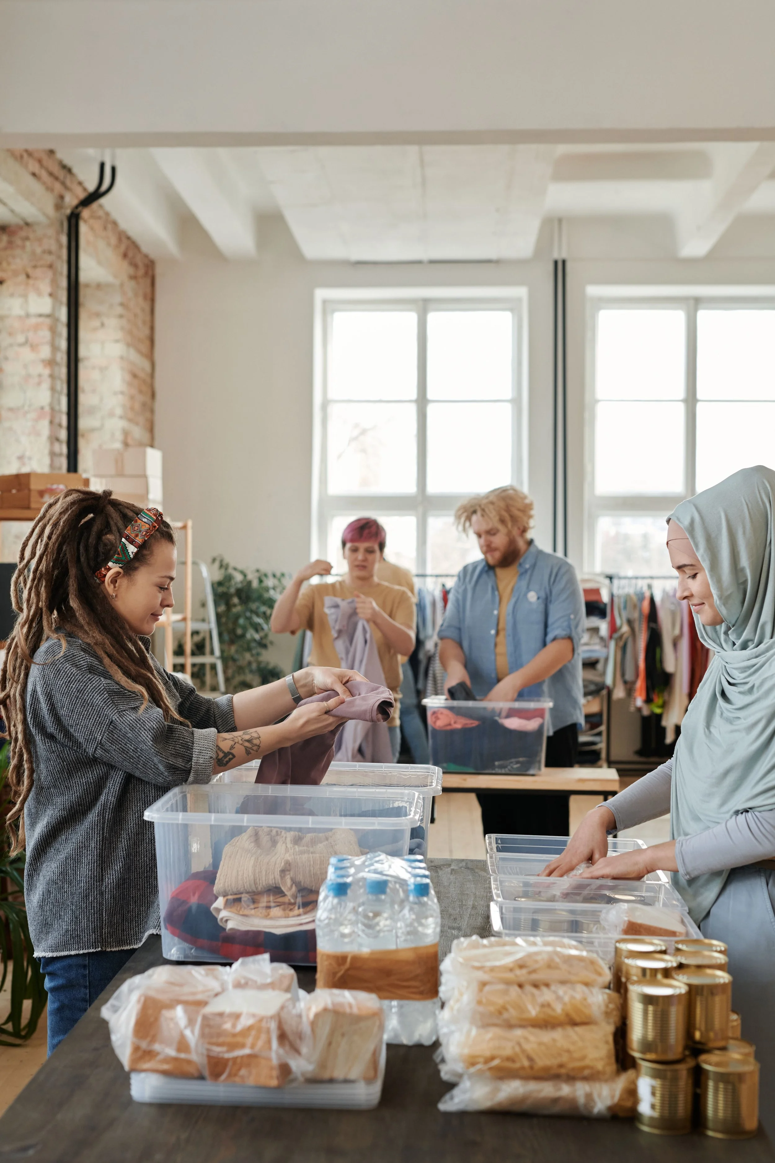 A group of people sorting and packing food and supplies in a bright, spacious room with large windows, exposed brick walls, and a high ceiling. The table in the foreground holds canned goods, packaged food, and water bottles.