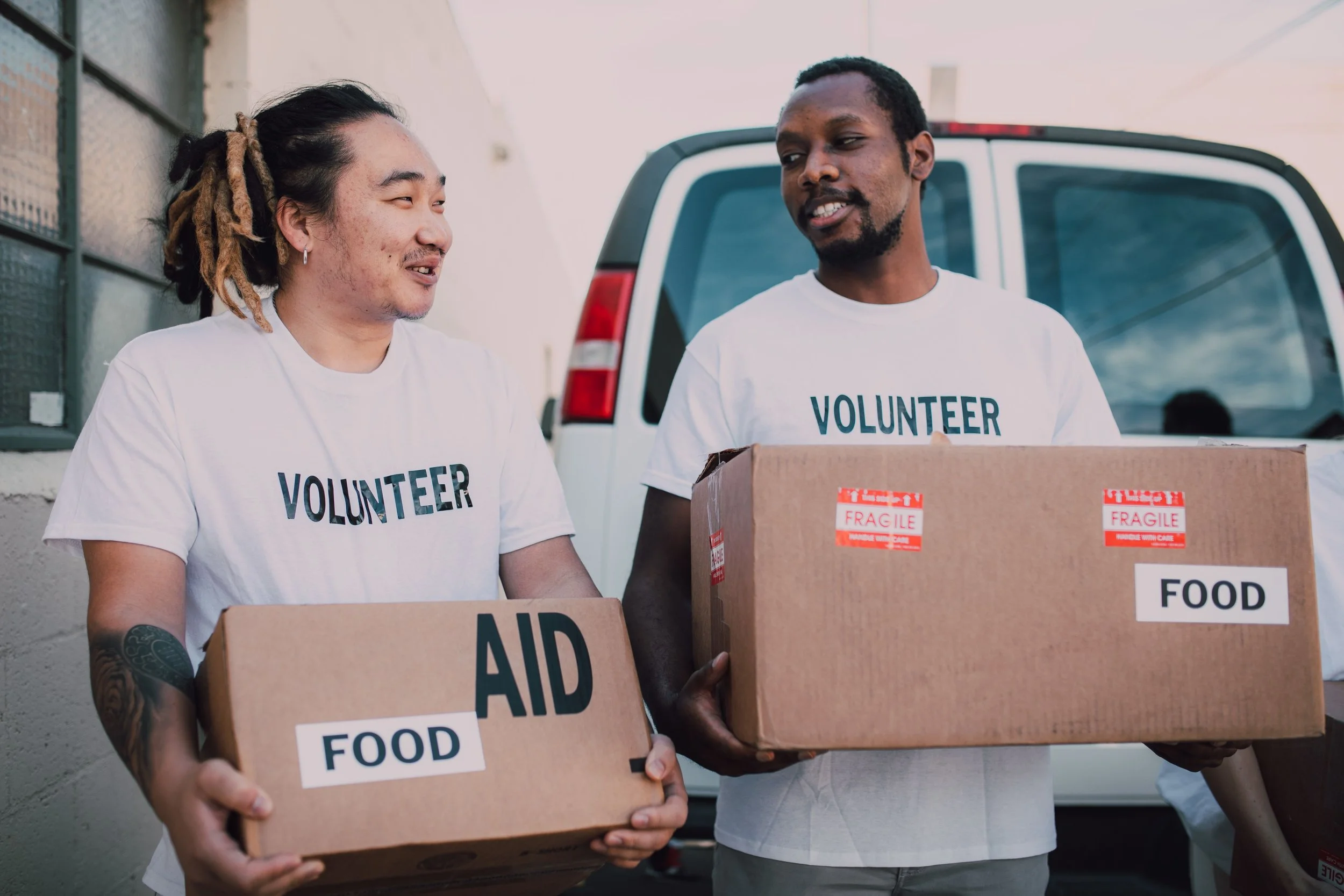 Two volunteers wearing white T-shirts with 'VOLUNTEER' printed on them are carrying cardboard boxes labeled 'FOOD' and 'AID.' They are standing outdoors in front of a vehicle.