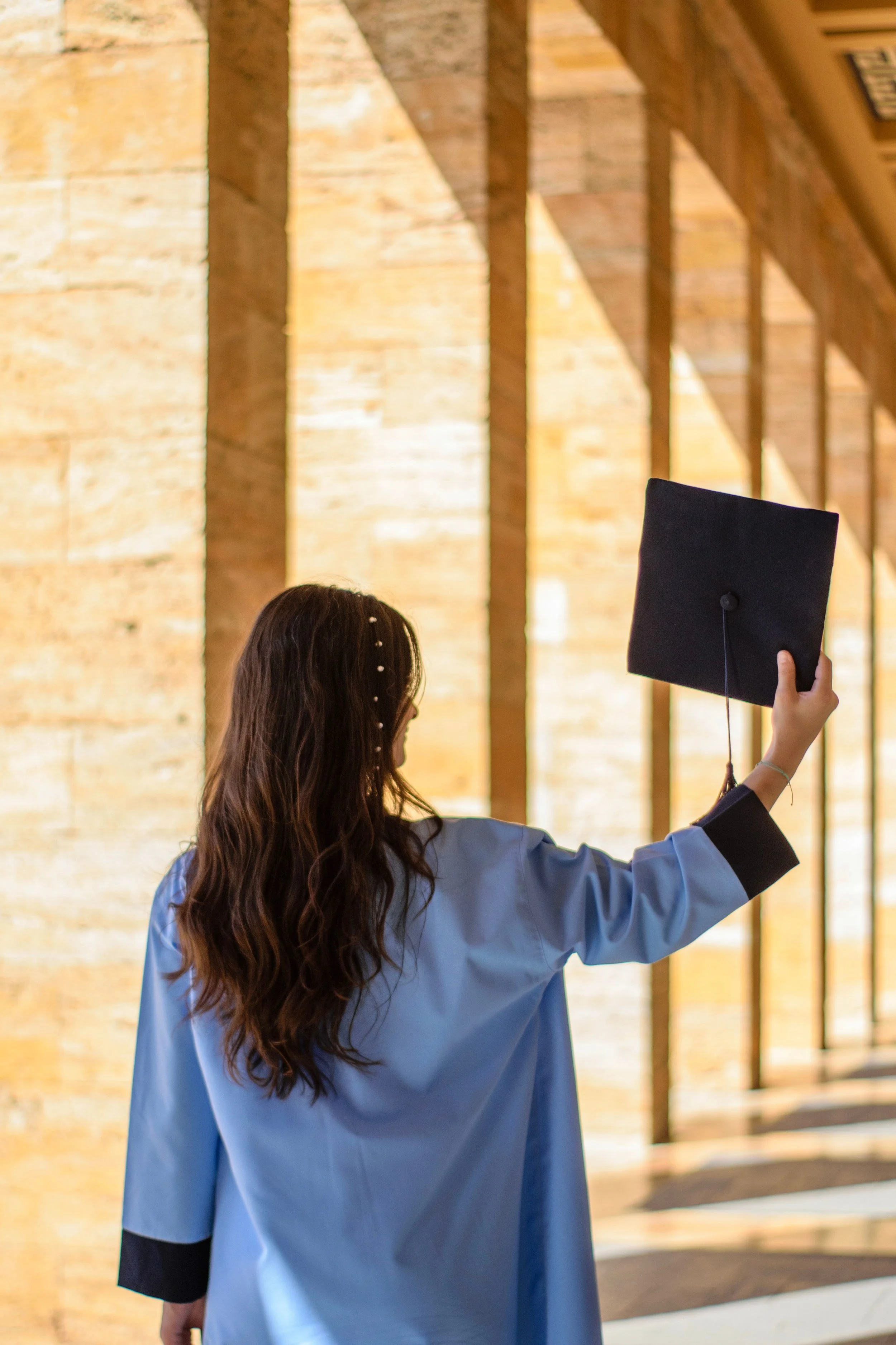 Young woman with long wavy brown hair wearing a blue graduation gown holding a black graduation cap with the tassel hanging down, standing indoors with a background of large brick and wood beams.
