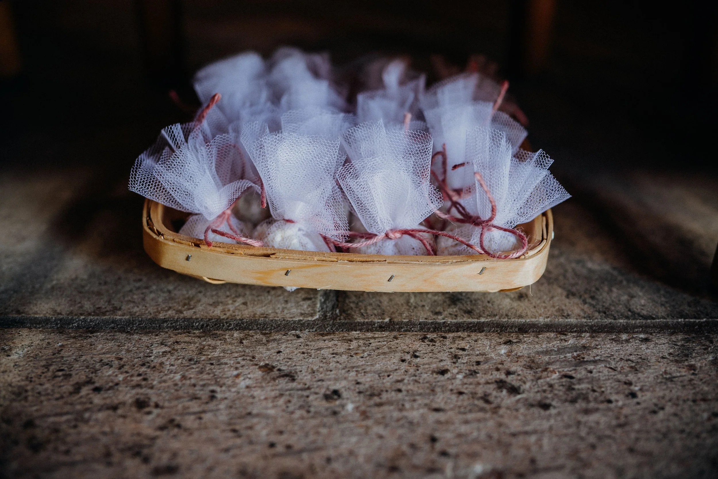 A basket containing multiple small items wrapped in white tulle fabric and tied with pink string, placed on a textured stone surface.